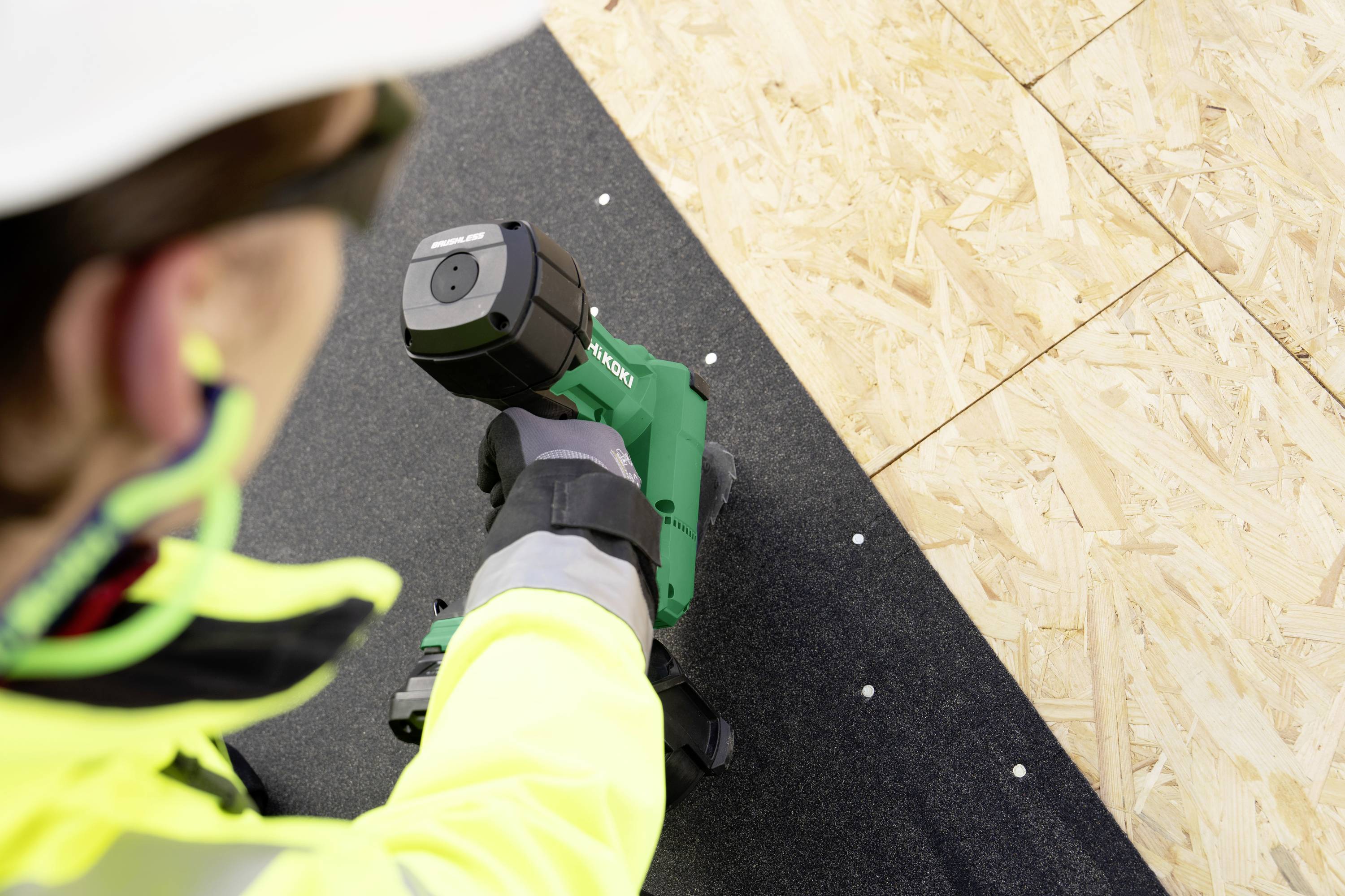 Ein Bauarbeiter in Schutzausrüstung verwendet einen grünen elektrischen Schraubendreher, um Holzplatten zu befestigen und dabei sichere Bauvorgehensweisen zu unterstreichen.