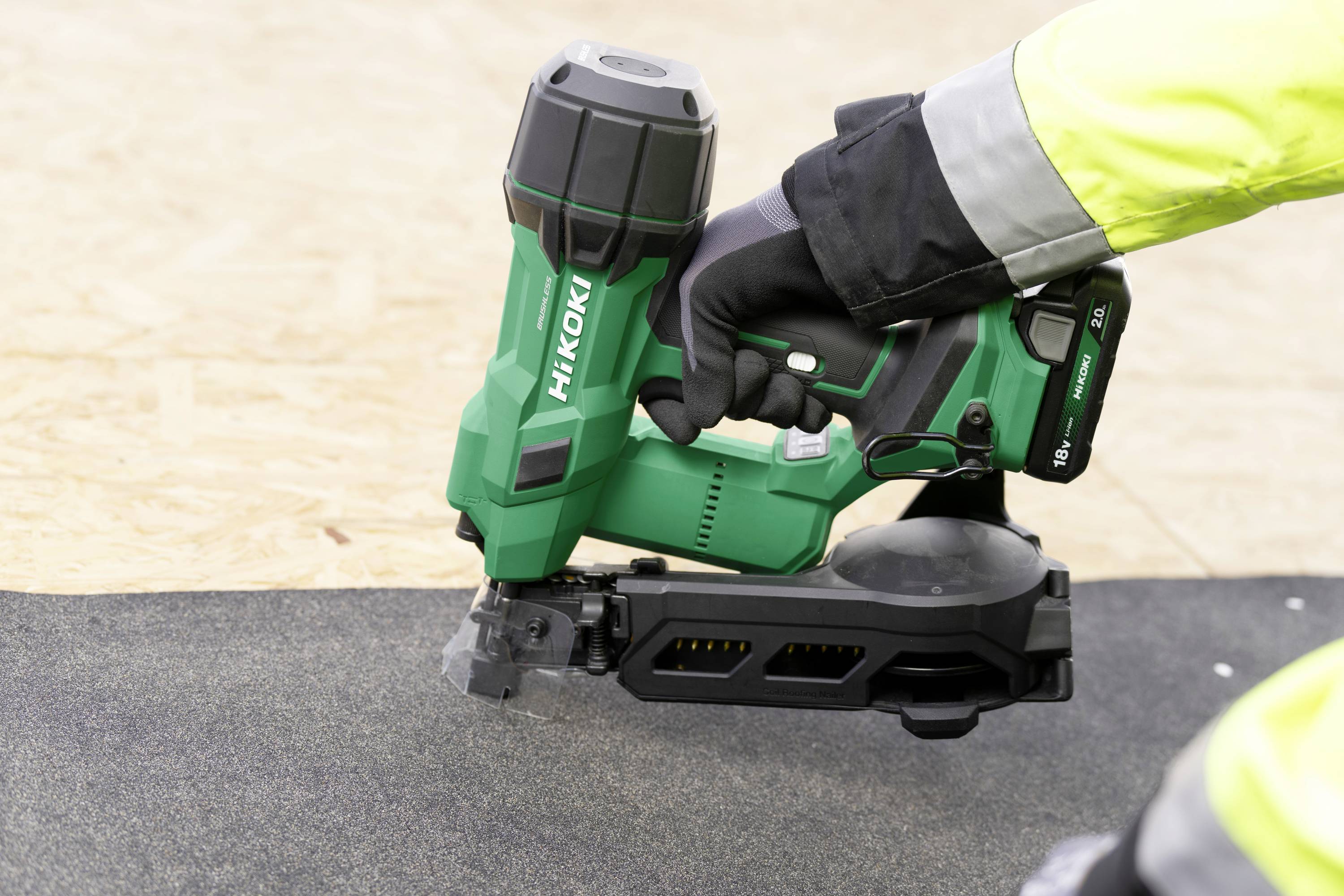 Eine Person in einer Warnweste verwendet einen grünen Hitachi-Nagler auf einer schwarzen Oberfläche, möglicherweise Dachdeckermaterial.