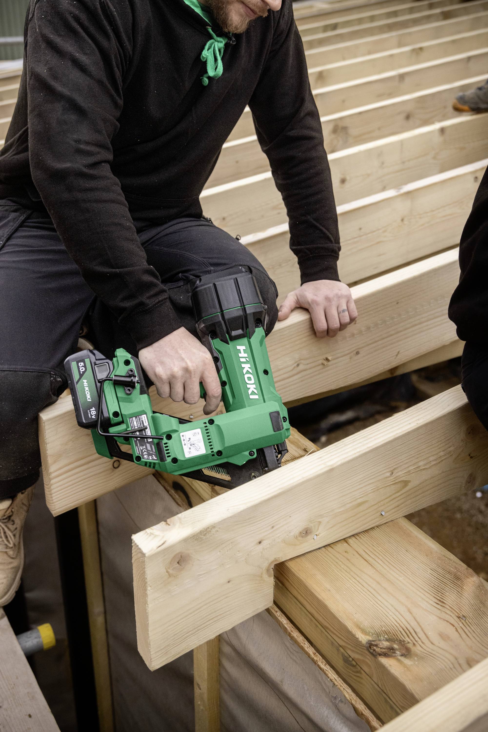 Eine Person verwendet ein grünes Elektrowerkzeug, um Holzbalken auf einer Baustelle zu befestigen und damit Zimmermanns- oder Bauarbeiten im Gange zu demonstrieren.