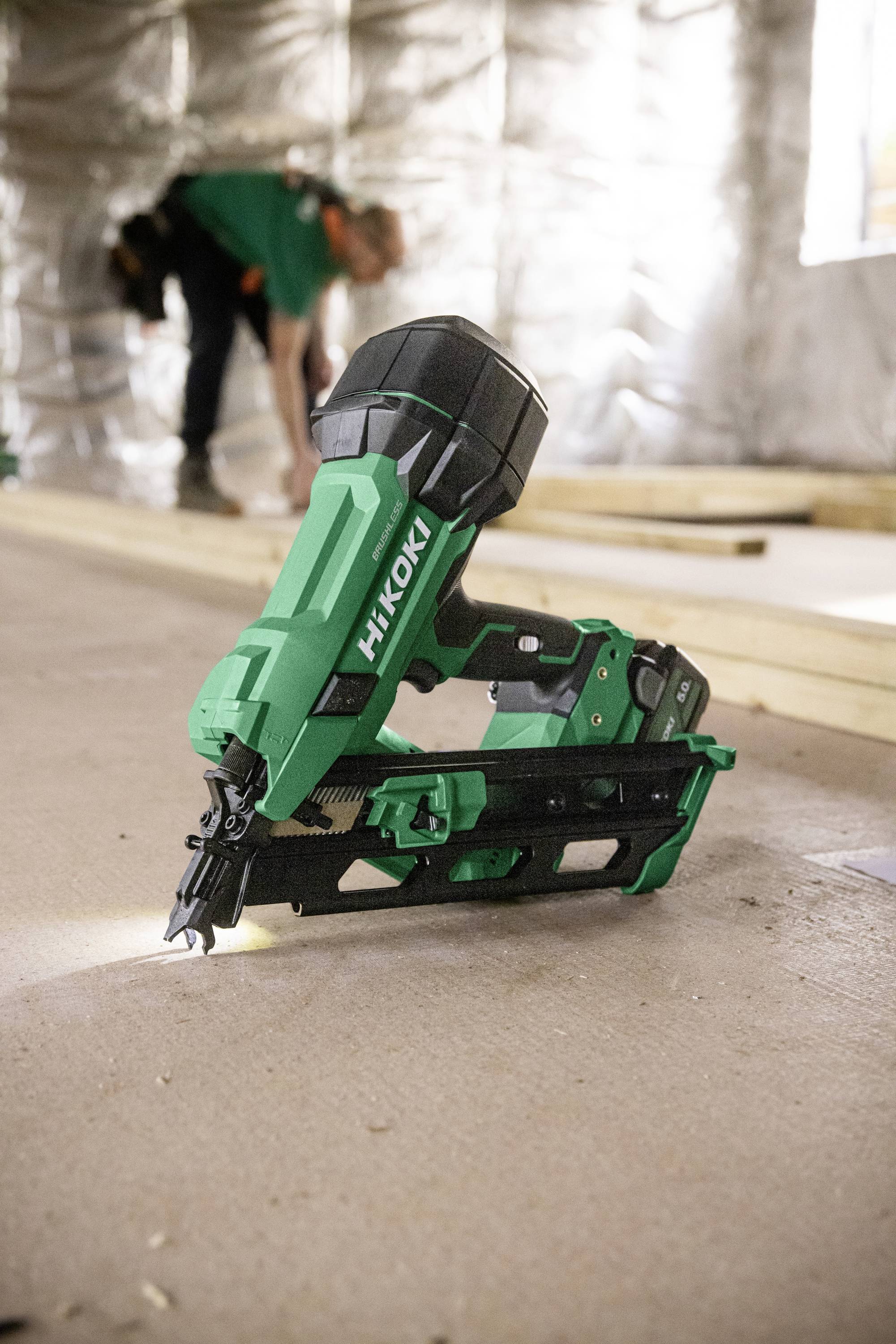 Eine grüne HiKOKI-Akku-Nagelmaschine ist positioniert, wobei Bauarbeiter und Holzbalken im unscharfen Hintergrund einer Baustelle sichtbar sind.