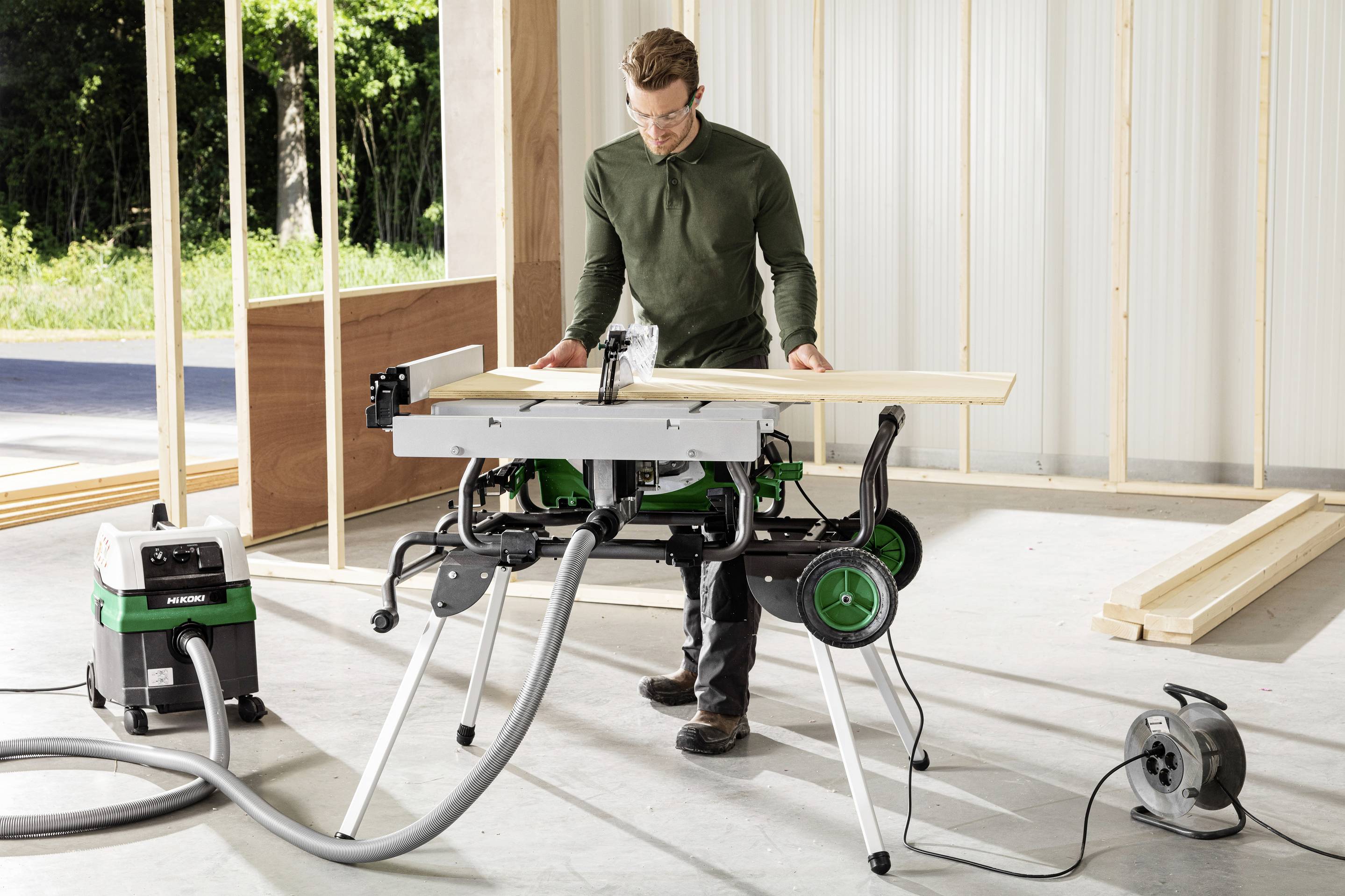 Eine Person steht in einem teilweise fertiggestellten Gebäude und verwendet eine Tischkreissäge, um Holzbretter zu schneiden. Bauwerkzeuge sind rundherum sichtbar.