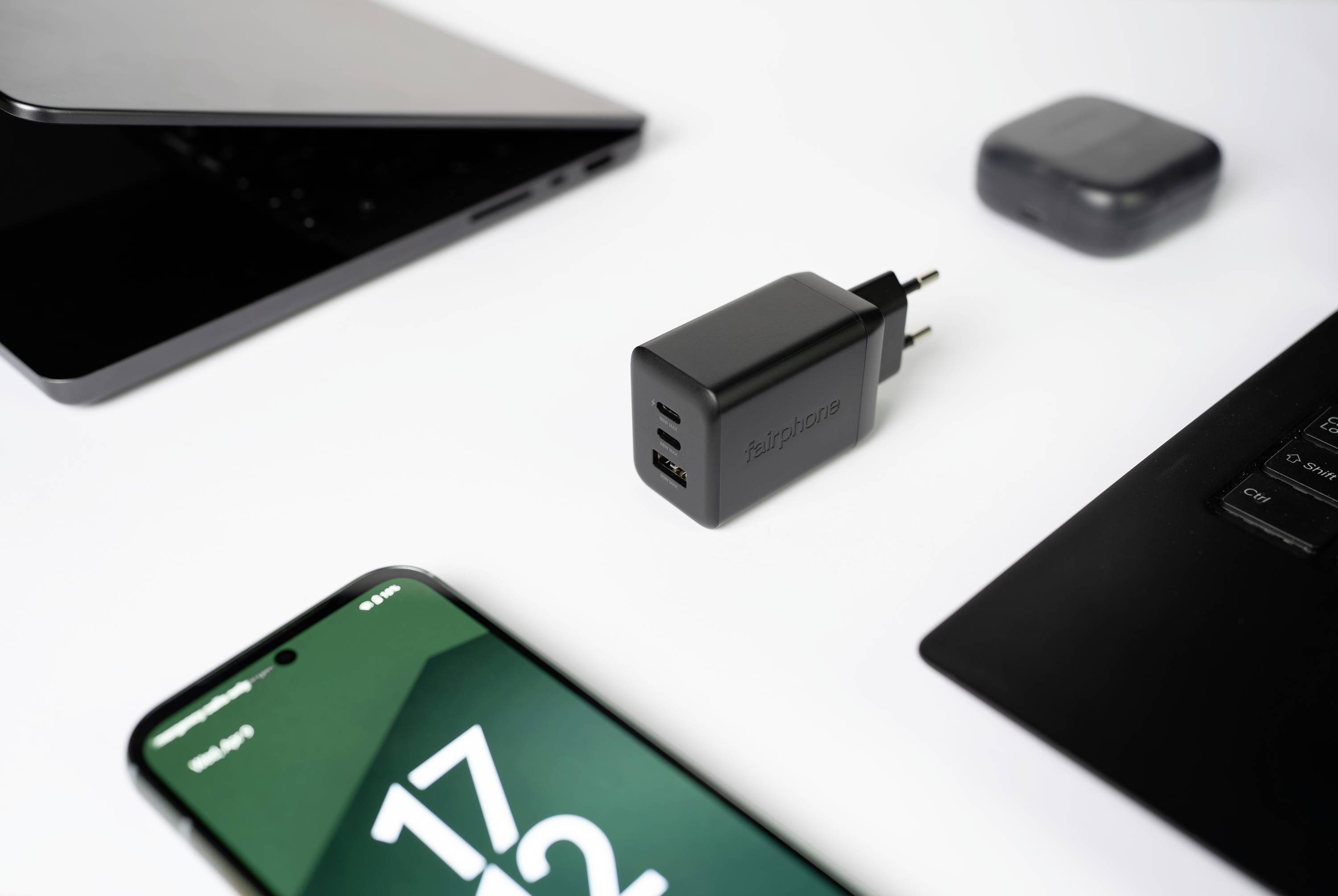 Ein schwarzer dualer USB-Wandladegerät, platziert zwischen einem Smartphone mit der Anzeige '17:23', einem halb geöffneten Laptop und drahtlosen Kopfhörern auf einer weißen Oberfläche.