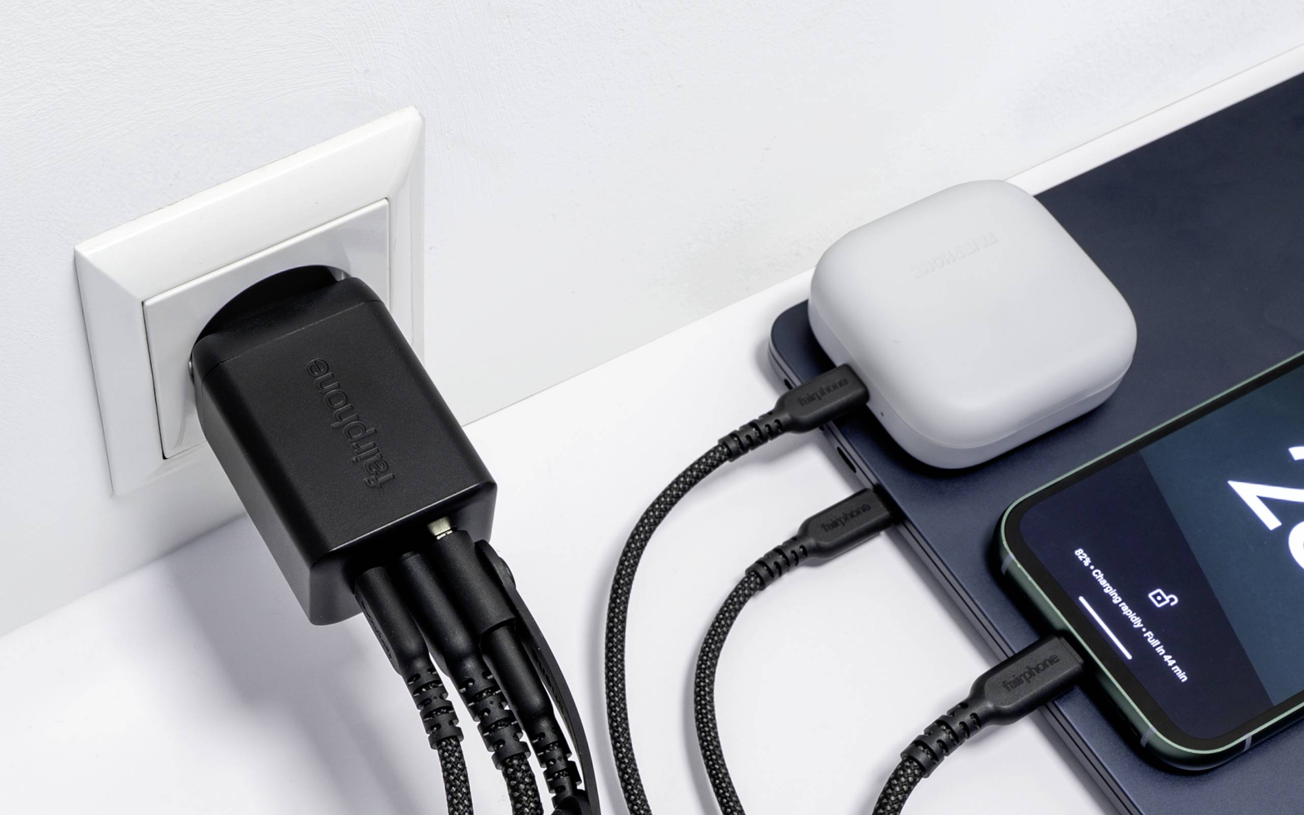 Eine Wandsteckdose mit einem Mehrfach-Ladegerät lädt gleichzeitig ein Smartphone, einen Laptop und ein Wireless-Ohrhörer-Etui und zeigt damit das gleichzeitige Aufladen mehrerer Geräte.