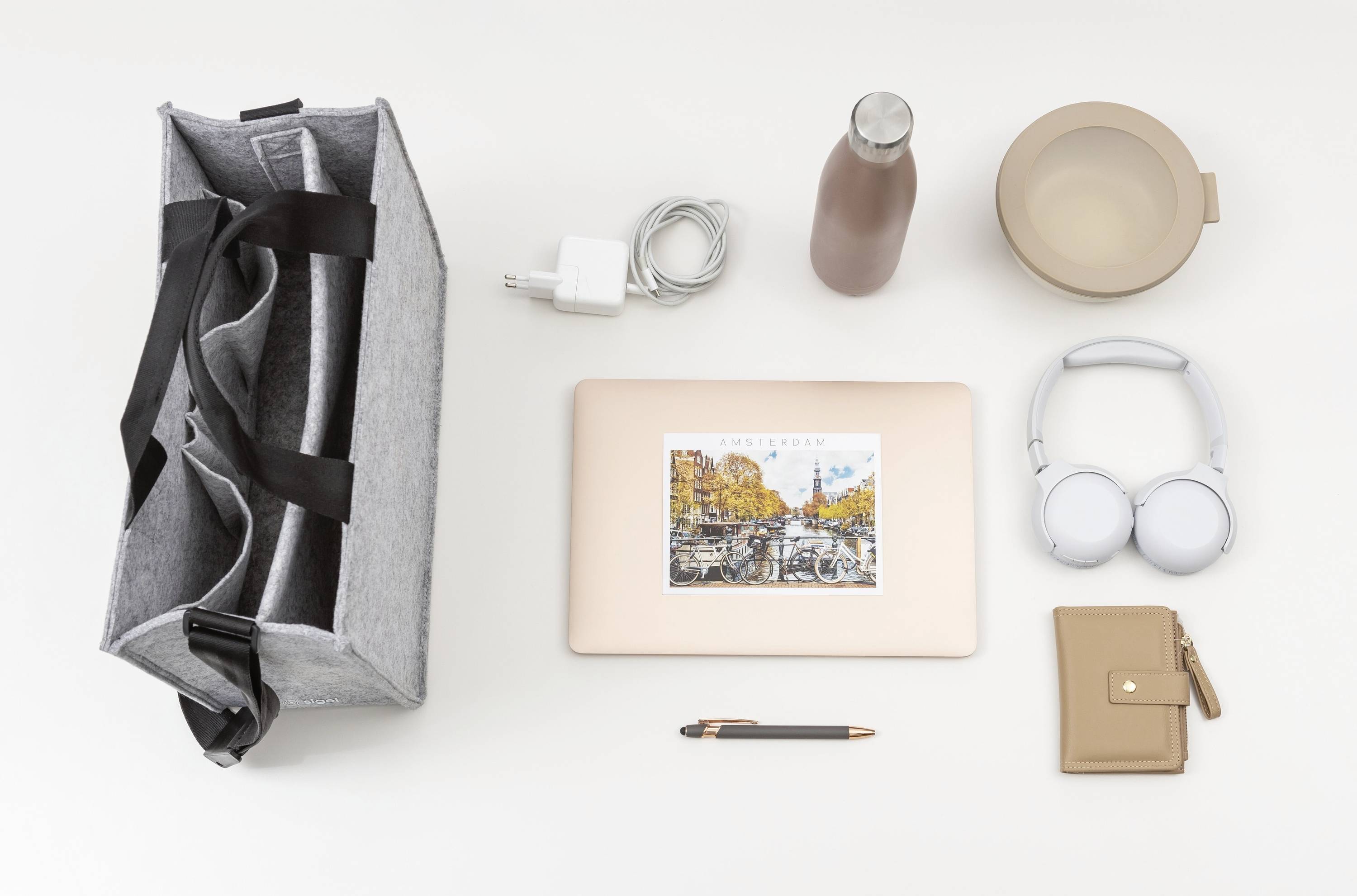 Tasche, Laptop mit Landschaftssticker, Kopfhörer, Geldbörse, Ladegerät, Wasserflasche, Schüssel und Stift ordentlich auf einem hellen Hintergrund angeordnet.