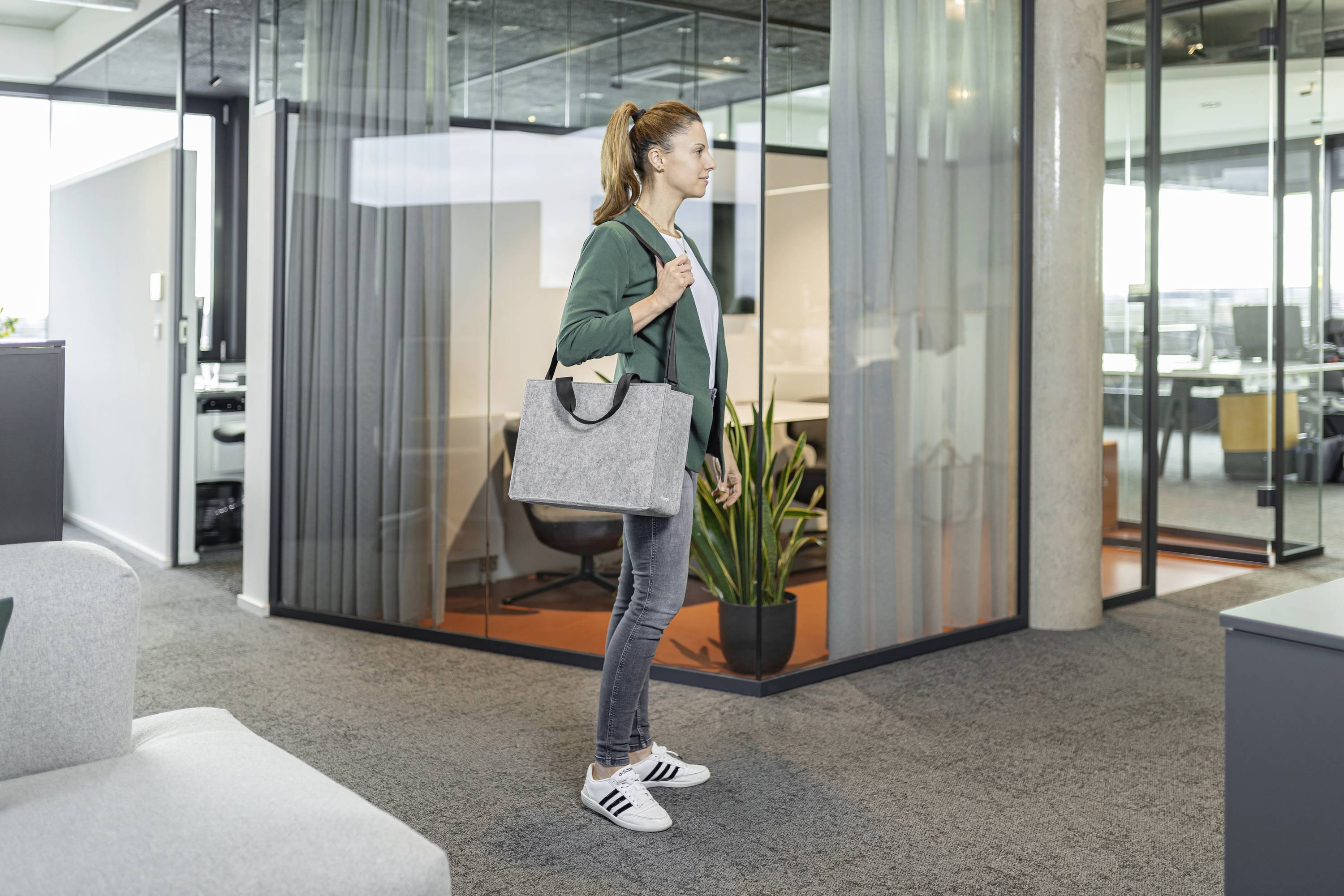 Eine Person, die in einem modernen Büro mit Glaswänden geht und eine graue Tasche trägt. Lässige Kleidung und minimalistische Einrichtung mit sichtbaren Pflanzen.