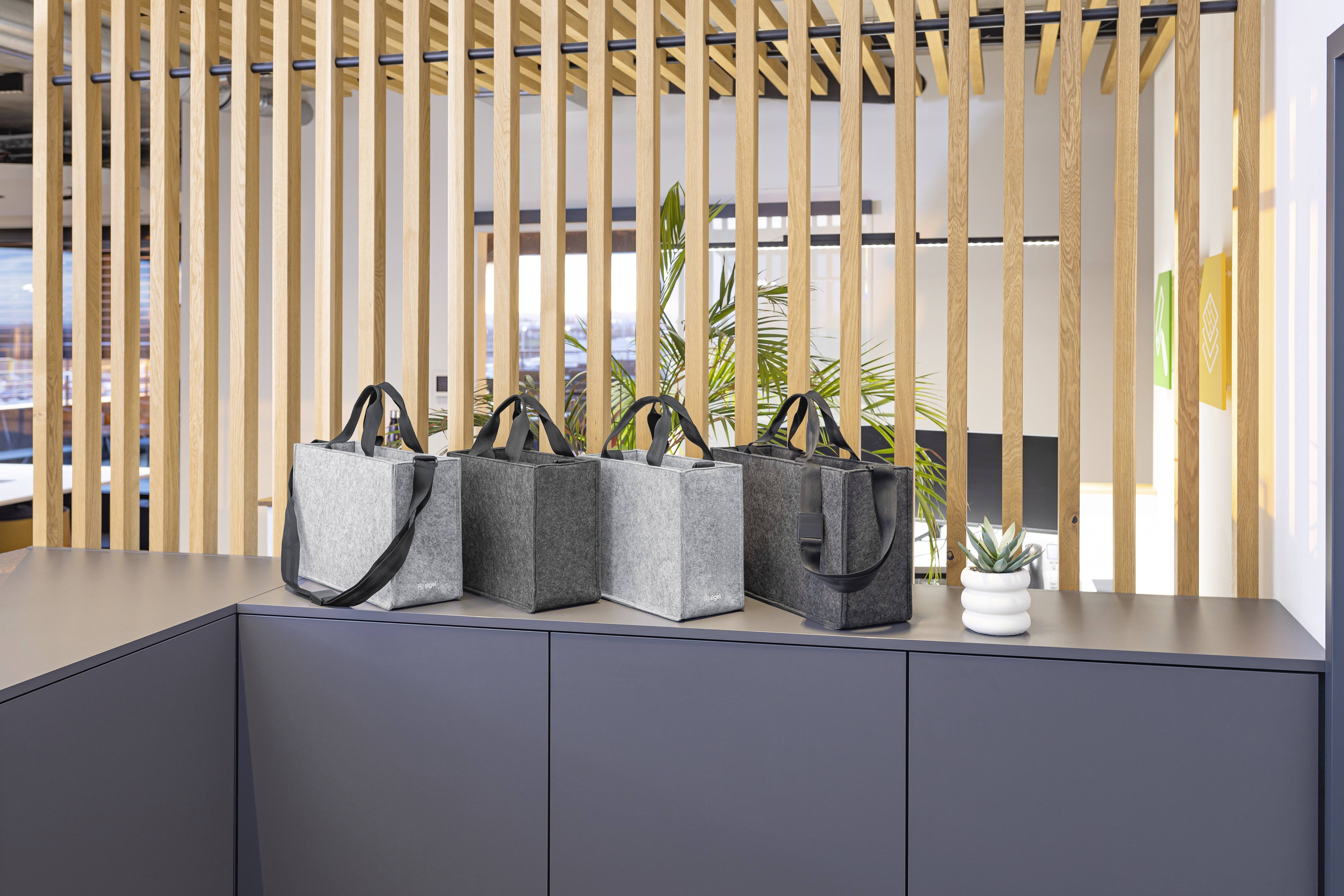 Vier graue Stofftaschen mit schwarzen Griffen werden auf einem grauen Schrank präsentiert. Eine Holzlamellenwand und eine Pflanze befinden sich im Hintergrund.