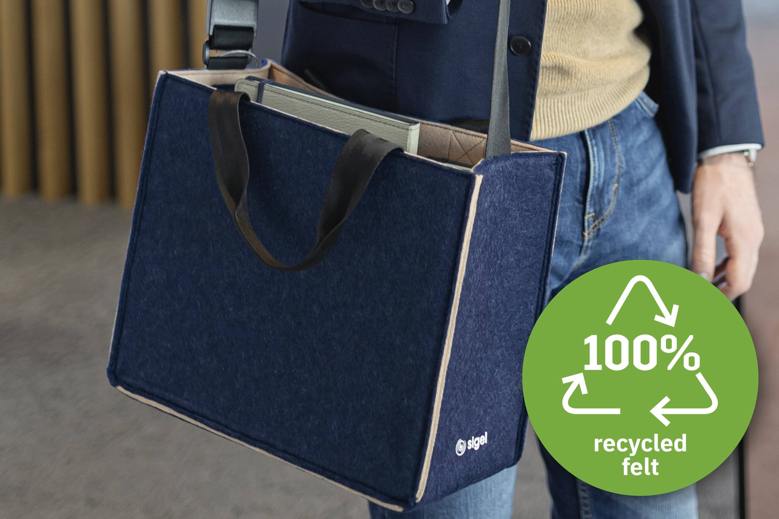 Eine Person trägt eine blaue Filztasche mit einem '100% recycelt' Symbol. Die Tasche wird zum Transport eines Laptops verwendet.