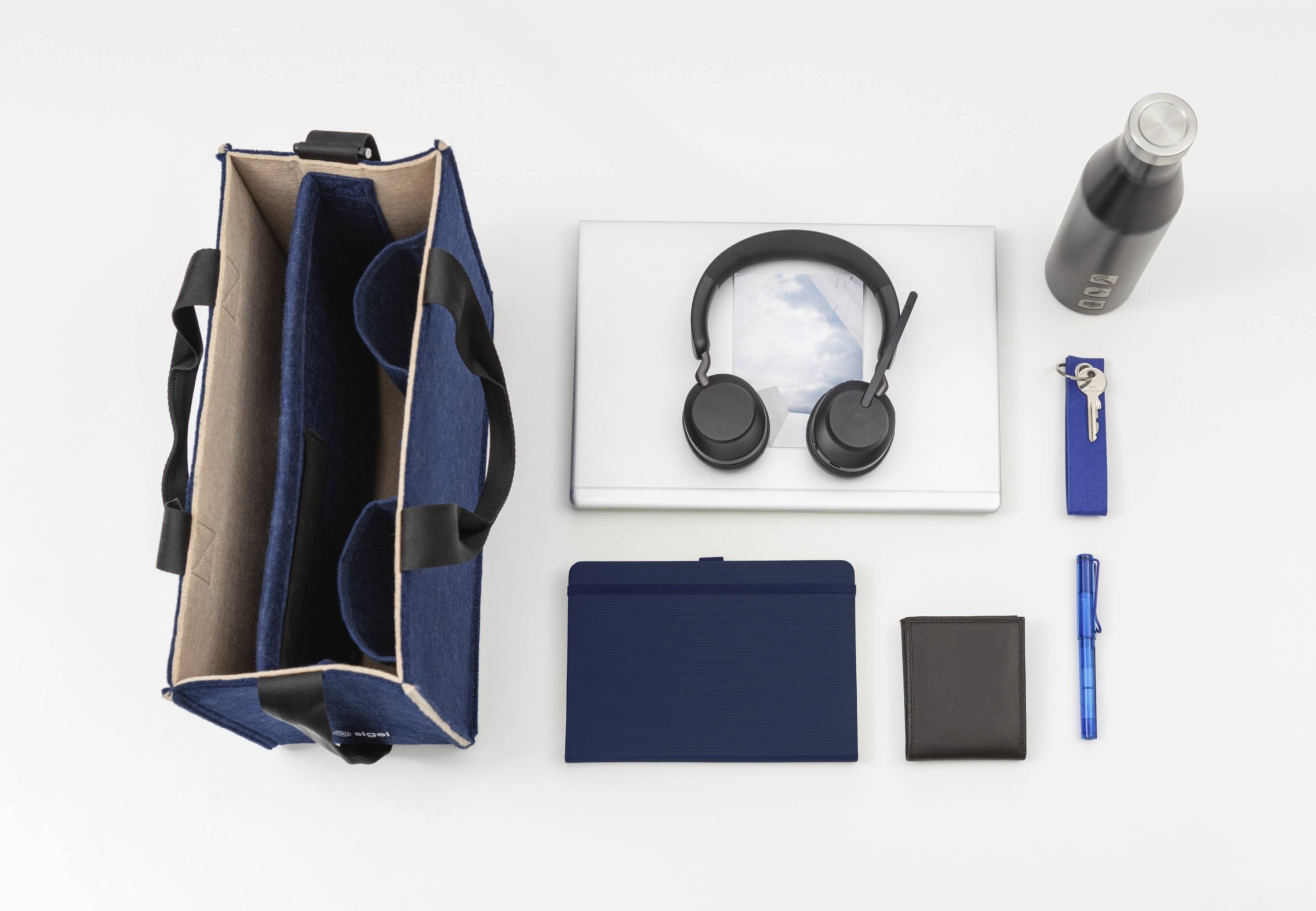 Eine sorgfältig angeordnete Anordnung auf einer weißen Oberfläche, die einen Laptop, Kopfhörer, eine Wasserflasche, eine Tragetasche, ein Notizbuch, eine Geldbörse, einen Stift und einen Schlüssel zeigt.