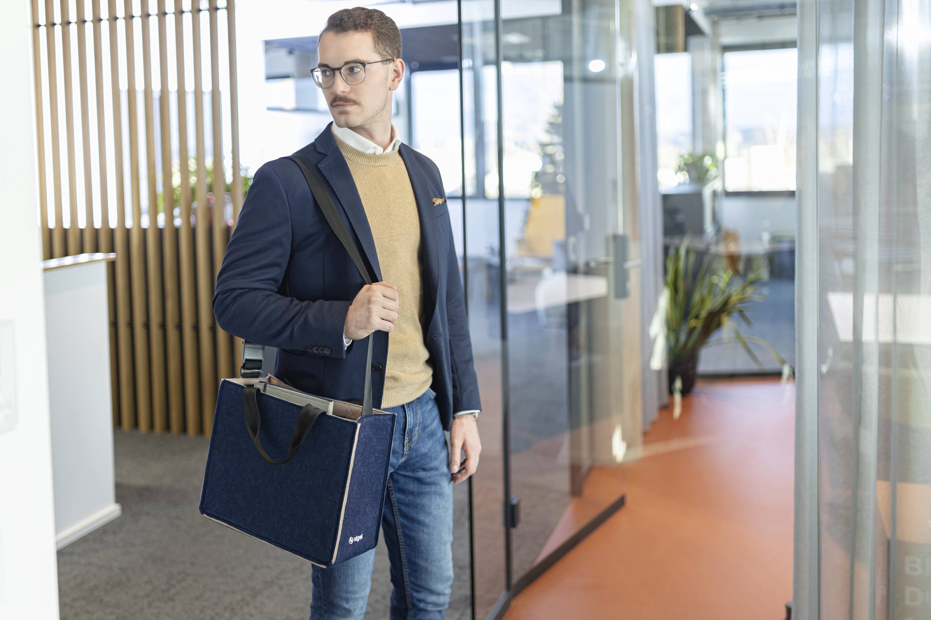 Ein Mann mit Brille, Blazer und Pullover trägt eine Laptoptasche und steht in einem modernen Büroraum mit Glaswänden.