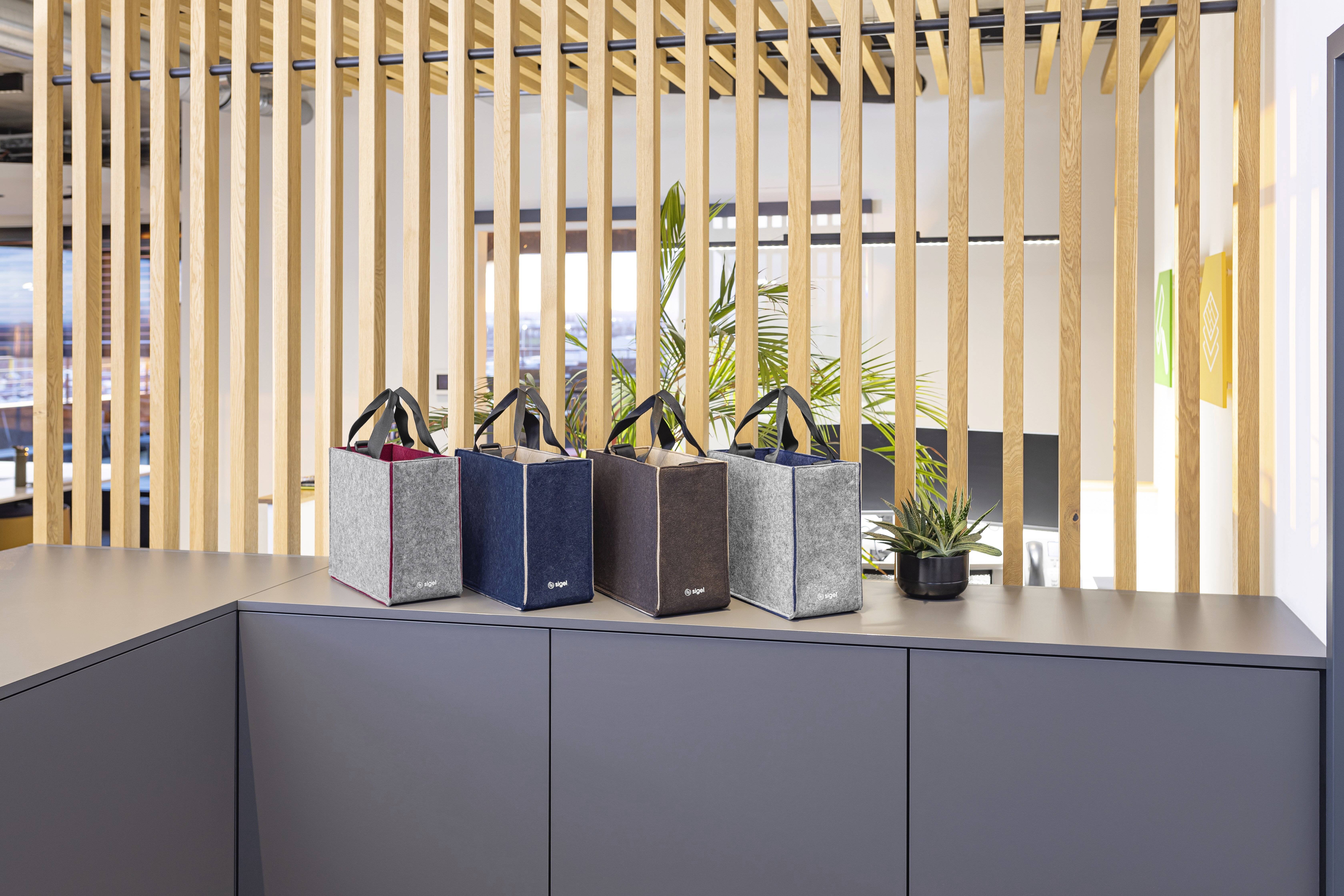 Vier Stofftaschen mit Griffen in verschiedenen Farben sind auf einer Theke aufgereiht, mit einer Holzlamellenwand und einer Topfpflanze im Hintergrund.