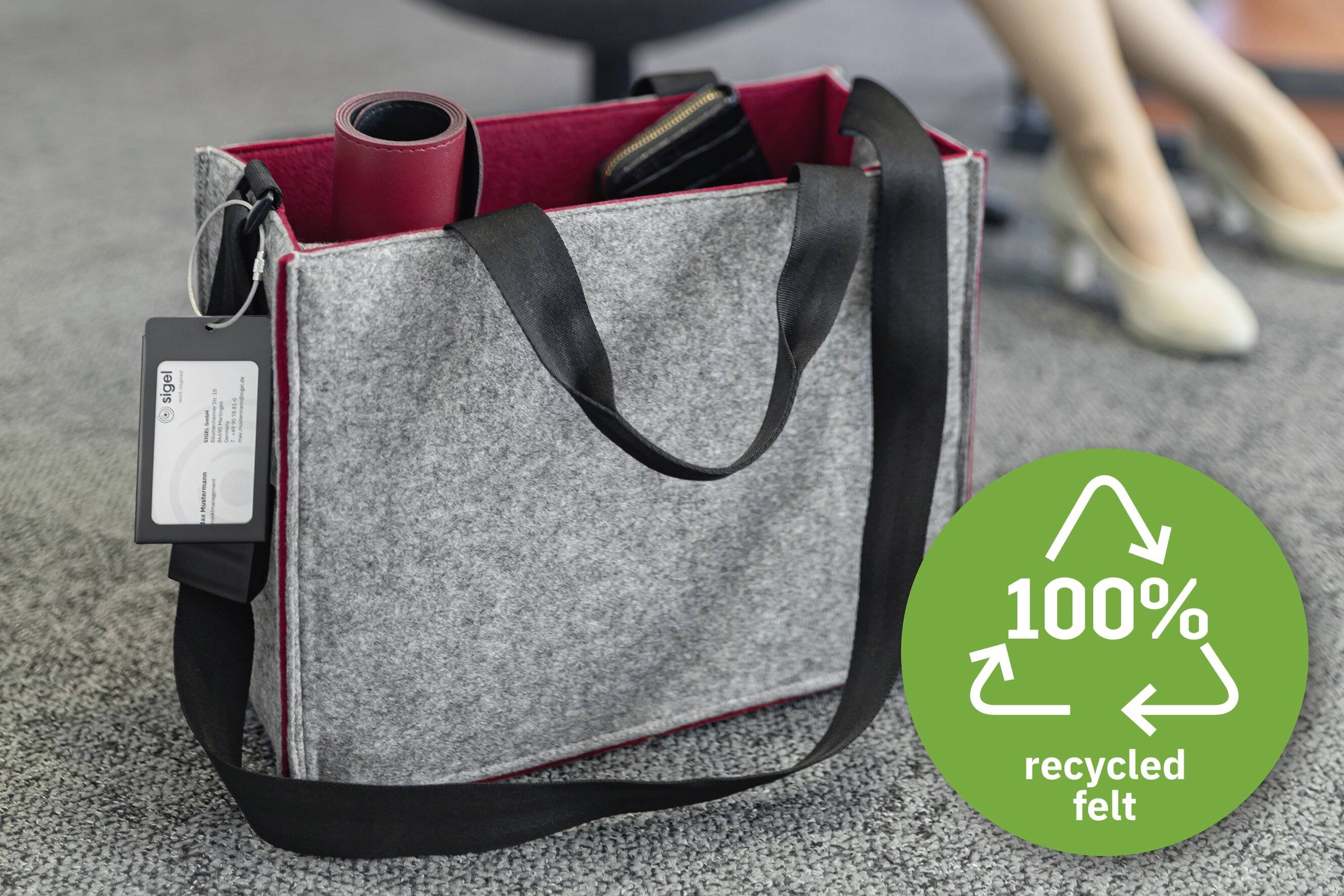 Eine graue Filz-Tragetasche mit schwarzen Henkeln steht auf einem Teppichboden. Sie enthält ein rot zusammengerolltes Objekt und eine Geldbörse. Ein Etikett mit der Aufschrift „100% recycelter Filz