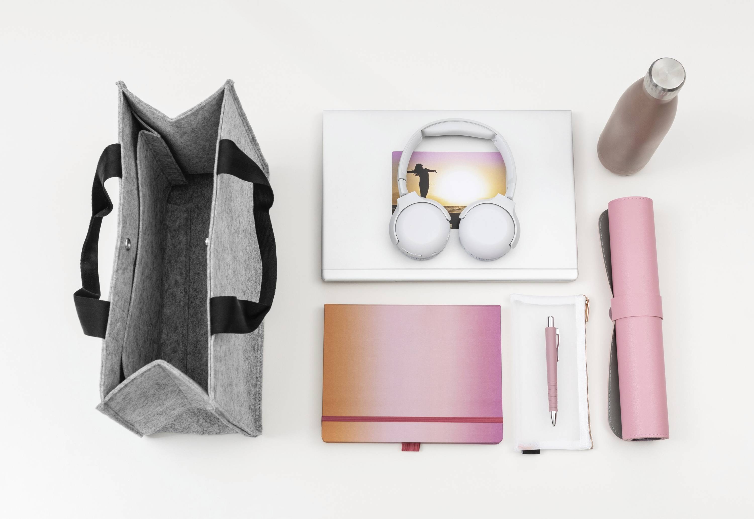 Graue Umhängetasche, Laptop mit Kopfhörern, rosa Wasserflasche, Notizbuch, Federmäppchen und rosa Matte, ordentlich auf einer weißen Oberfläche angeordnet.