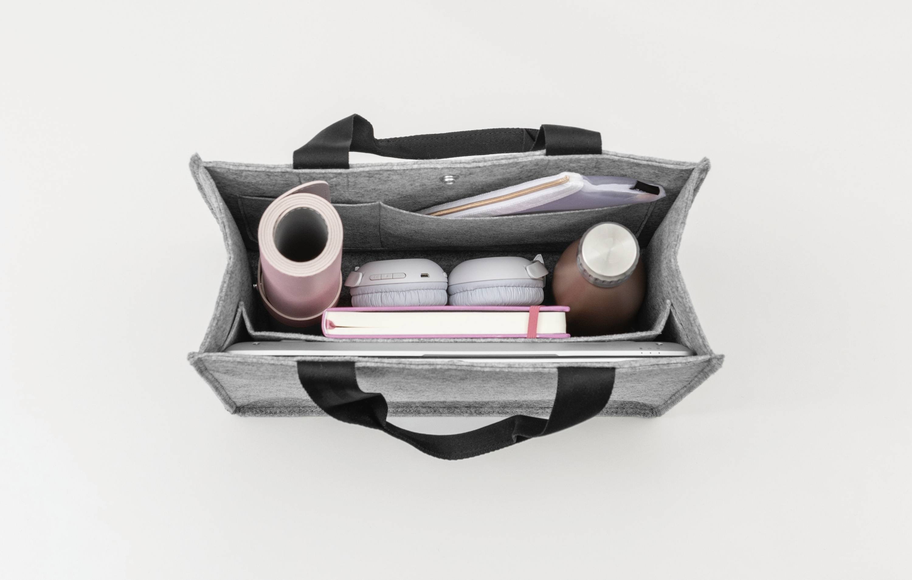 Draufsicht einer geöffneten grauen Tasche vor weißem Hintergrund, die eine pinke Wasserflasche, Kopfhörer, ein Notizbuch und eine braune Flasche enthält.