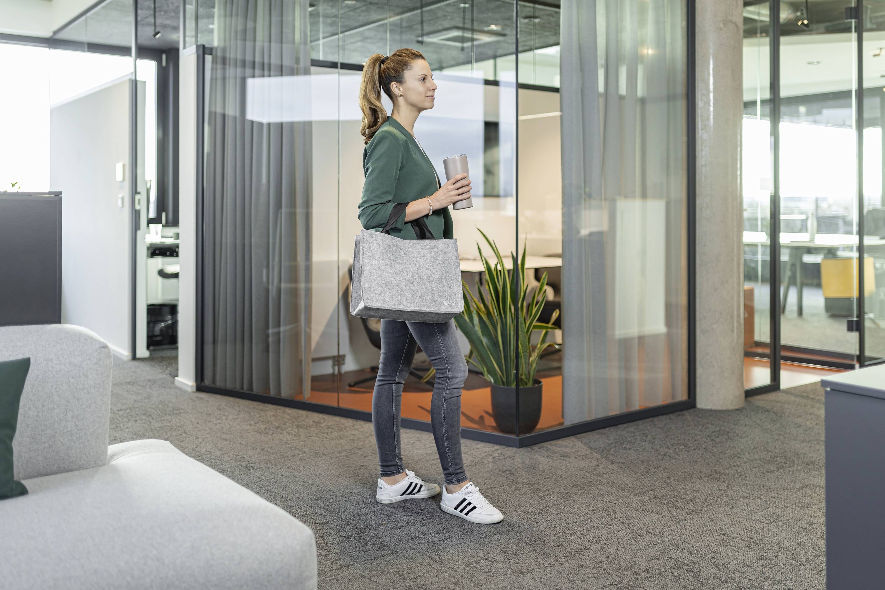 Eine Frau in Jeans und einer grünen Jacke geht durch ein modernes Büro und trägt einen Thermobecher und eine graue Tasche, mit Glaswänden und einer Pflanze in der Nähe.