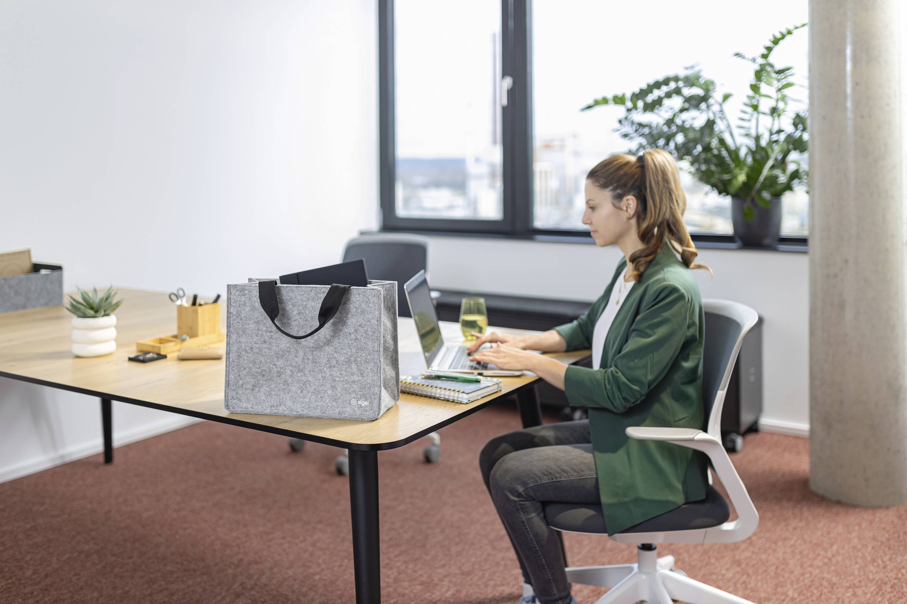 Eine Frau sitzt an einem Schreibtisch und tippt auf einem Laptop in einem modernen Büro mit einem großen Fenster, Pflanzen und einer Filztasche auf dem Schreibtisch.