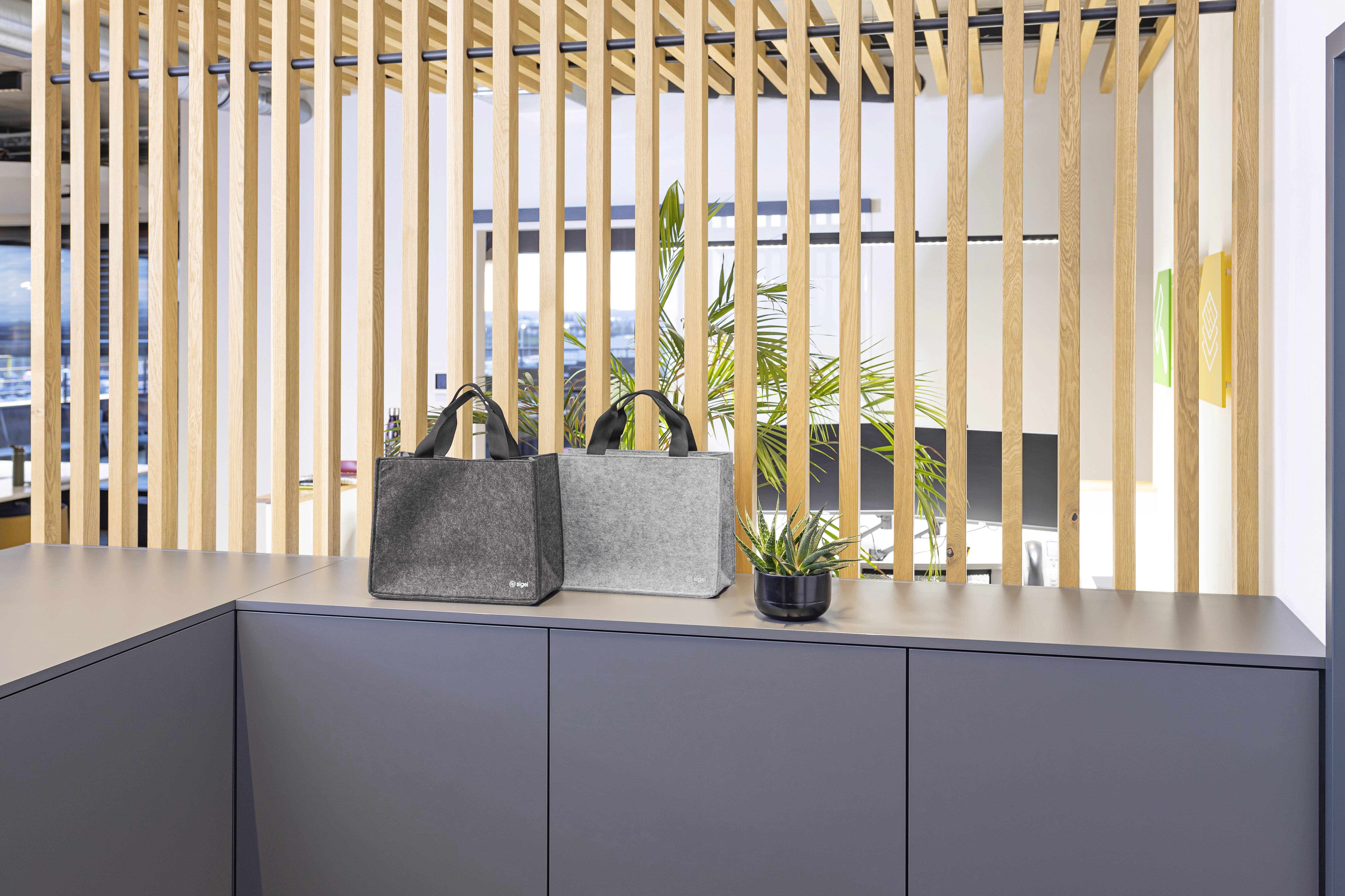 Zwei graue Tragetaschen auf einer modernen grauen Arbeitsplatte, vor einem Hintergrund aus Holzlatten und einer grünen Pflanze, was eine minimalistische Ästhetik erzeugt.