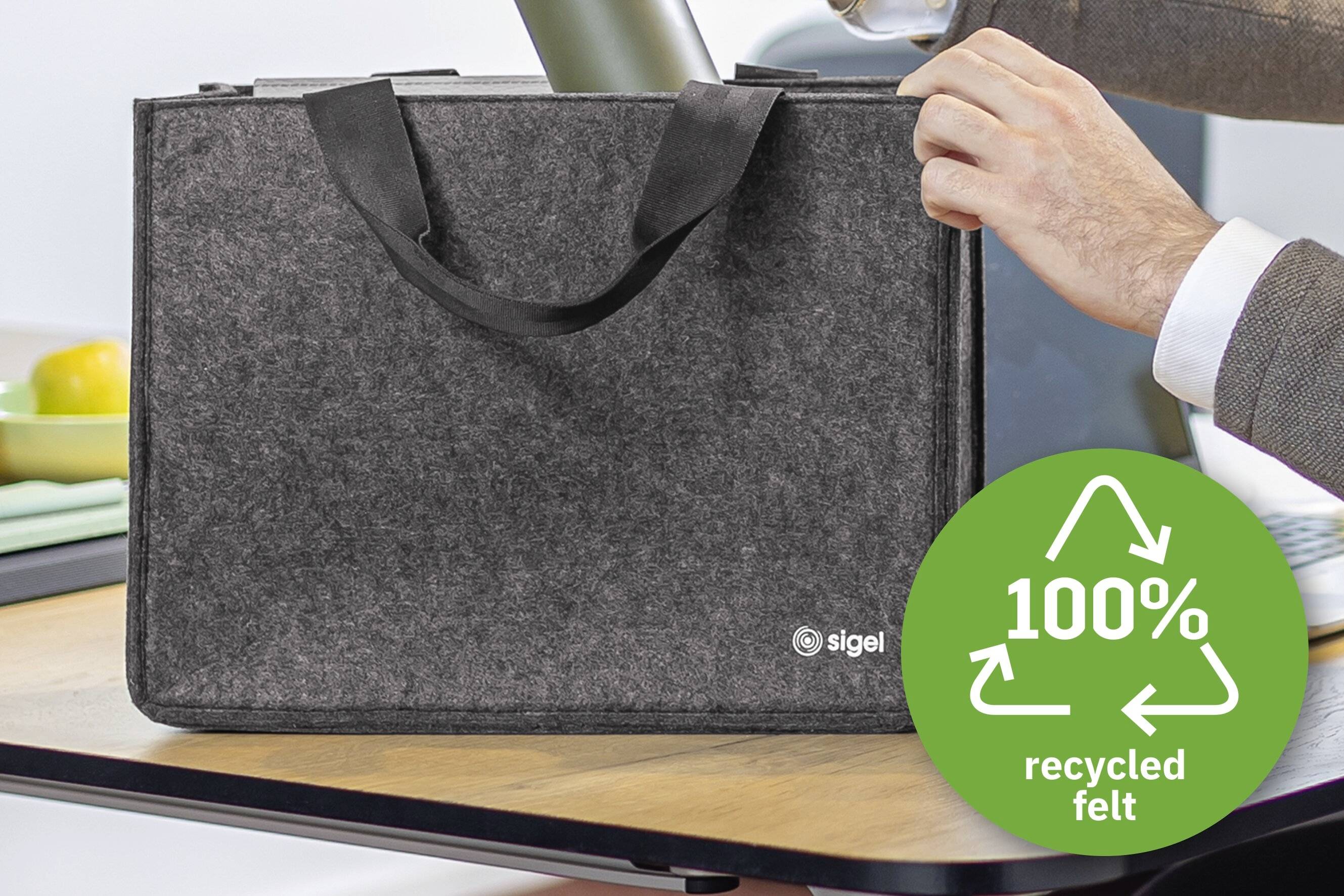Eine Person hält eine graue Filztasche auf einem Tisch. Die Tasche hat zwei Henkel und ein Logo. Ein grünes Abzeichen zeigt '100% recycelter Filz' mit Pfeilen, die das Recycling symbolisieren.
