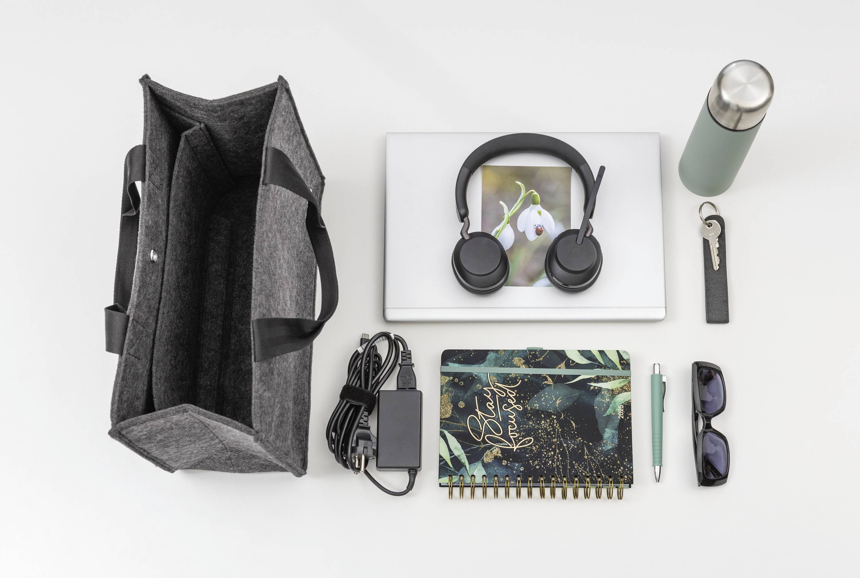 Eine ordentlich angeordnete Sammlung täglicher Gegenstände: eine graue Tragetasche, Laptop, Kopfhörer, Notizbuch, Wasserflasche, Ladegerät, Sonnenbrille, Stift und Schlüsselanhänger auf einer weißen Oberfläche.