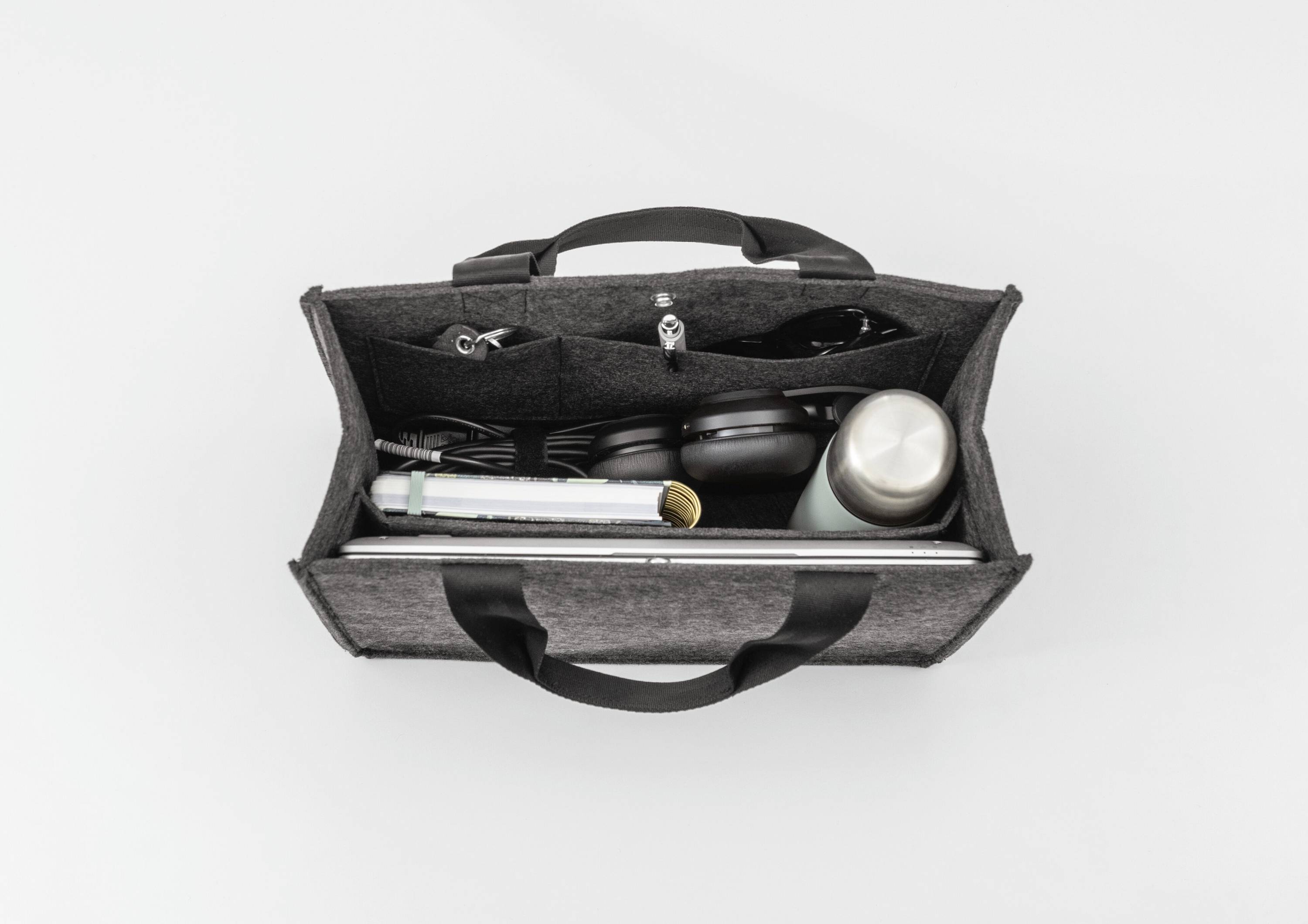 Eine Draufsicht einer geöffneten grauen Tragetasche mit schwarzen Griffen, die einen Laptop, Kopfhörer, einen Thermobecher und verschiedene kleine Gegenstände enthält.