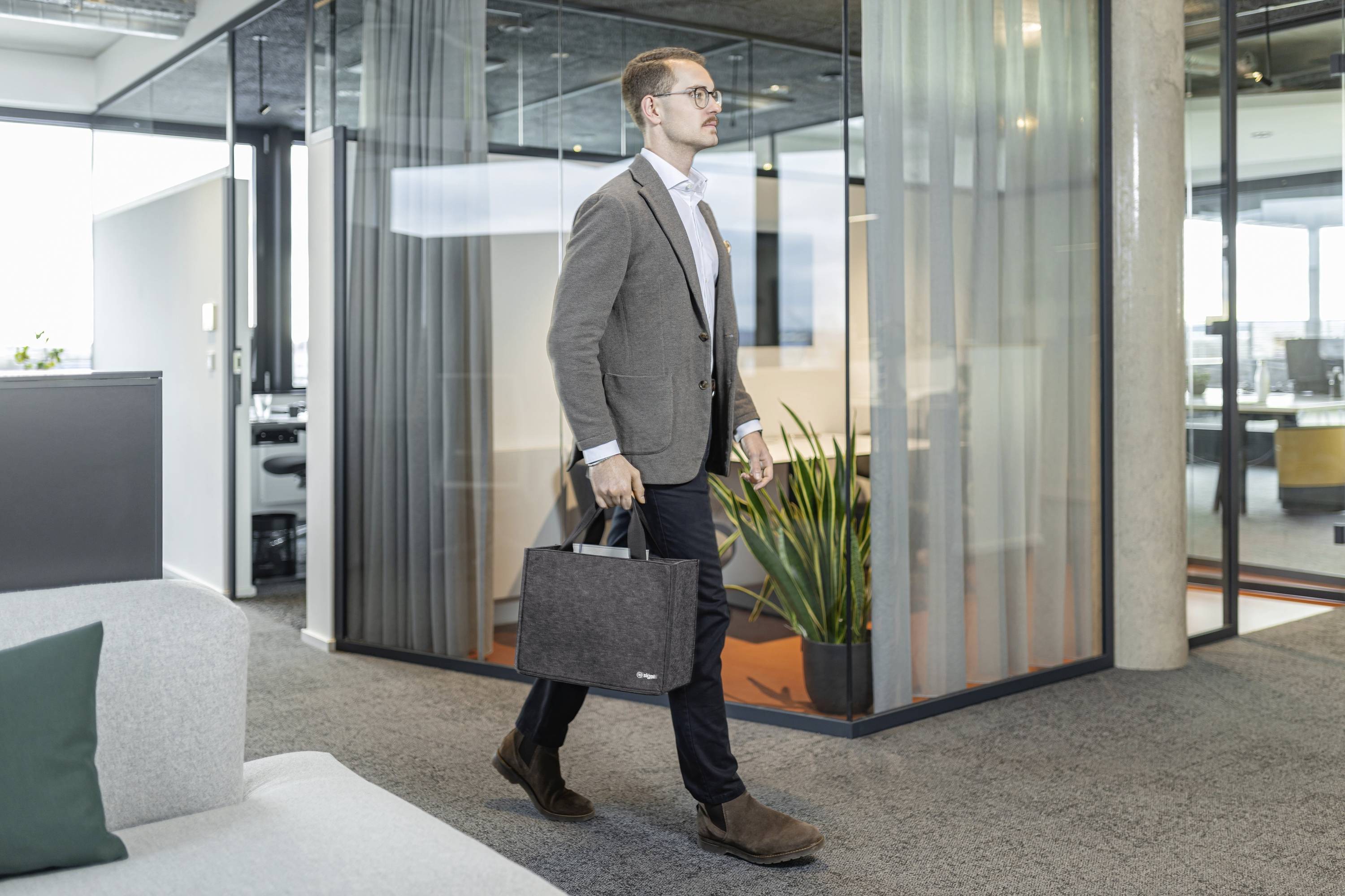 Ein Mann in Geschäftskleidung geht durch ein modernes Büro mit Glastrennwänden und trägt eine Laptoptasche, was eine professionelle Arbeitsumgebung vermittelt.