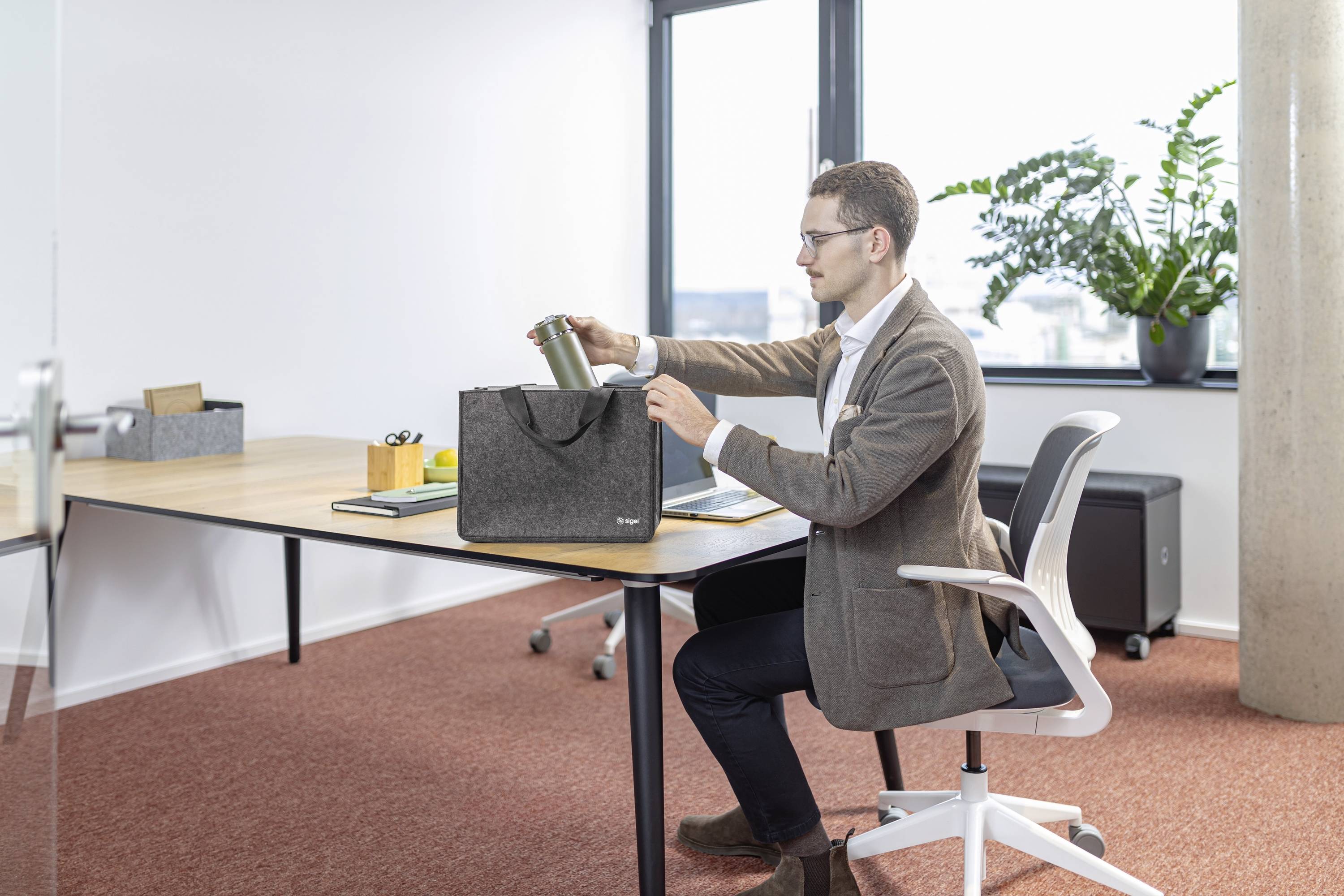 Eine Person sitzt an einem Schreibtisch in einem Büro und legt einen Thermobecher in eine graue Tasche. Der Raum hat ein großes Fenster, eine Zimmerpflanze und Büroausrüstung.