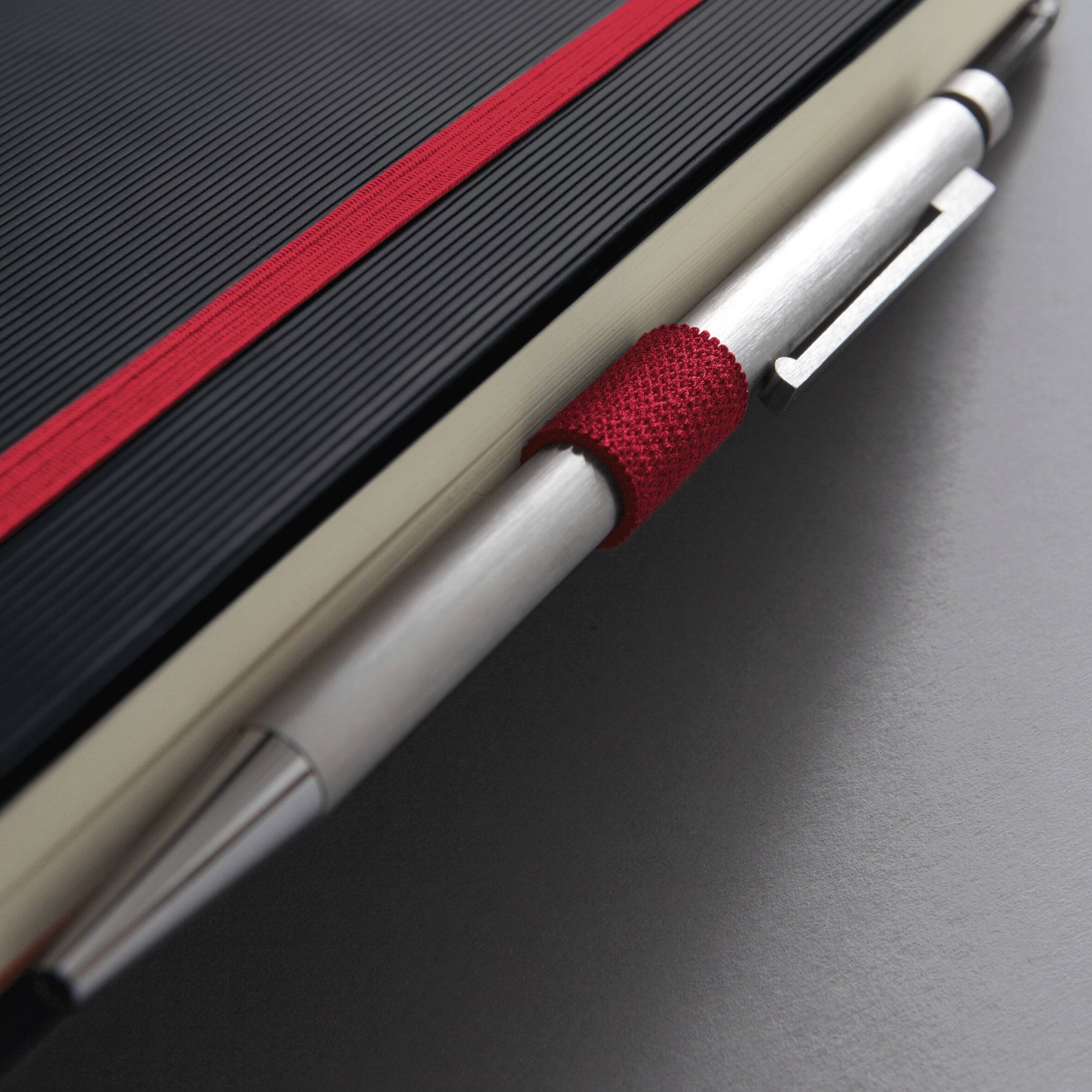 Ein geschlossenes schwarzes Notizbuch mit einem roten Gummiband, versehen mit einem silbernen Stift, der an der Seite eingeklemmt ist und auf einer grauen Oberfläche liegt.