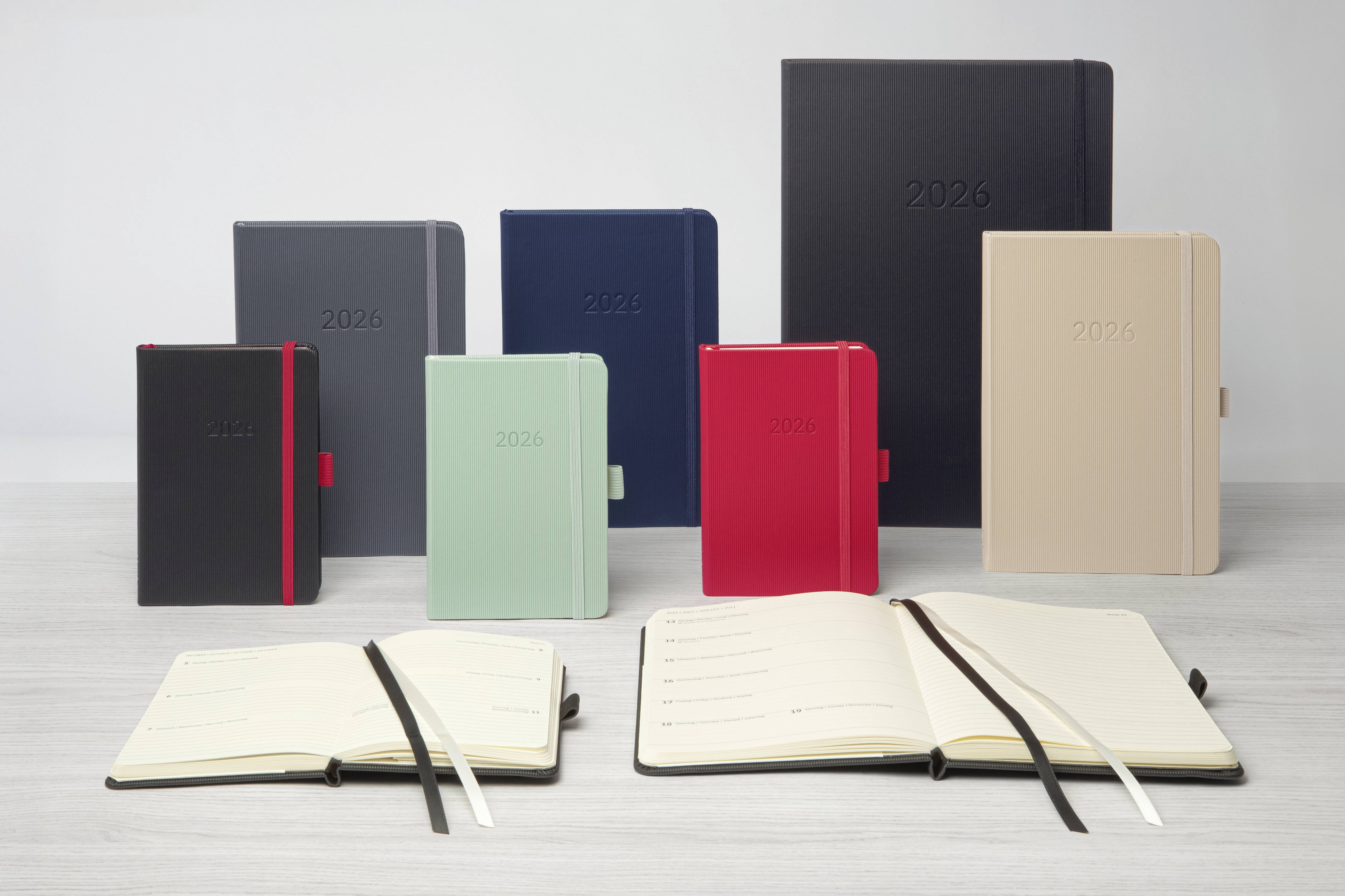 Eine Sammlung von Planern für das Jahr 2026 in verschiedenen Farben und Größen, wobei einige geöffnet sind und Linienblätter zeigen, ausgelegt auf einer Holzoberfläche.