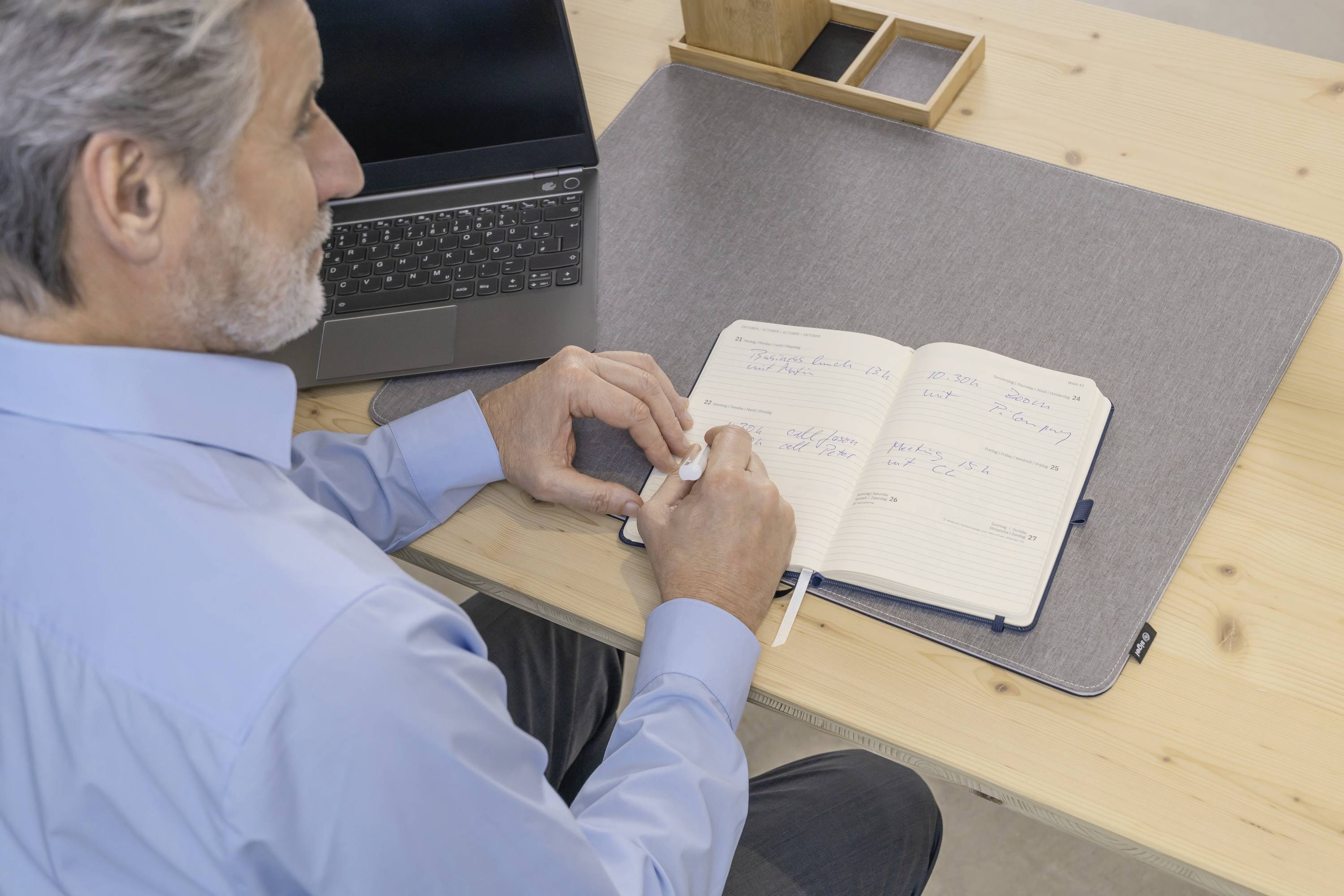 Eine Person in einem blauen Hemd macht Notizen in einem Notizbuch an einem Schreibtisch mit einem Laptop und stellt eine Arbeitsplatzszene dar.