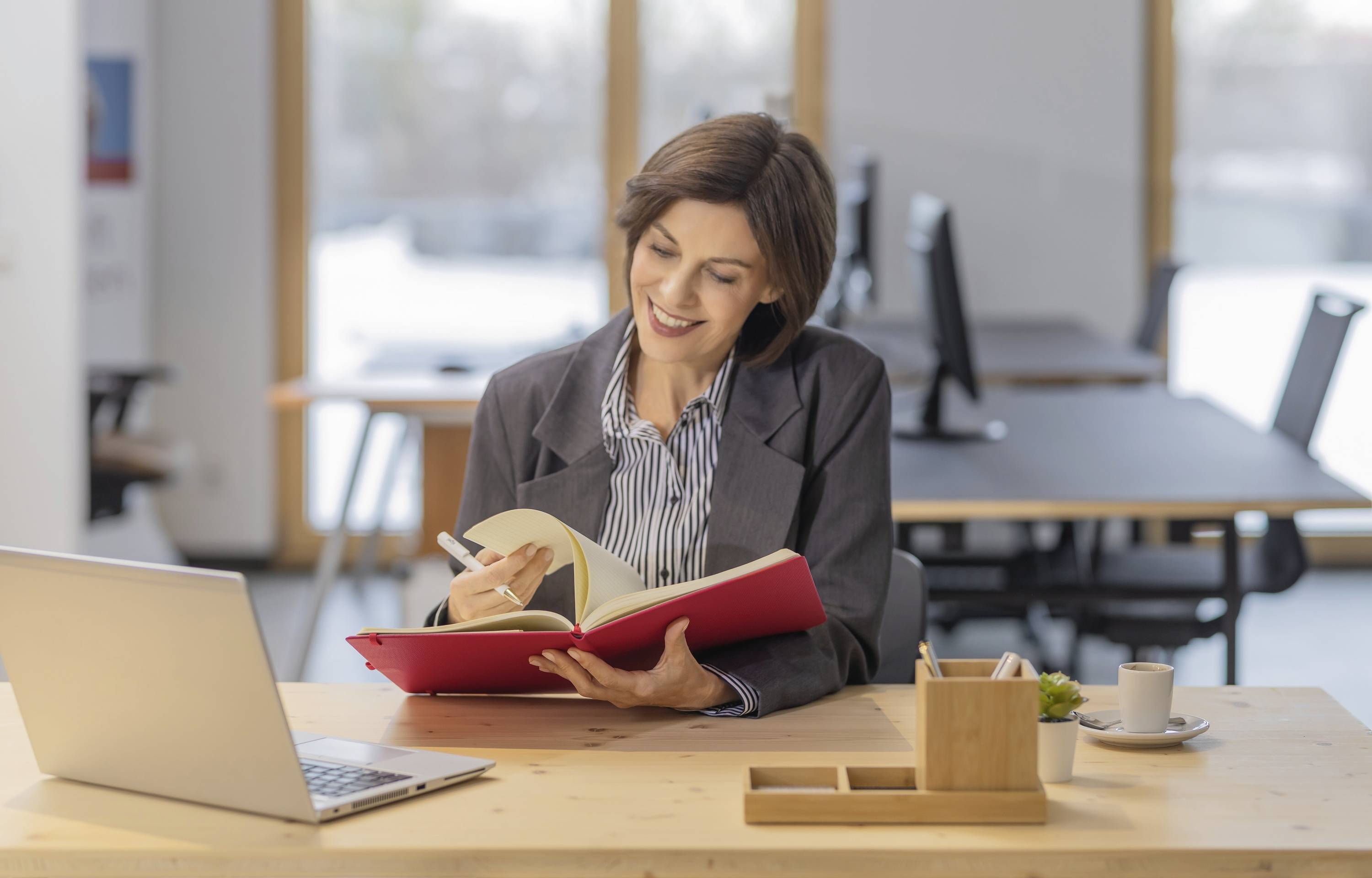 Eine Frau in einem Anzug sitzt an einem Schreibtisch in einem modernen Büro, lächelt und schreibt in ein rotes Notizbuch, mit einem Laptop und Kaffee in der Nähe.