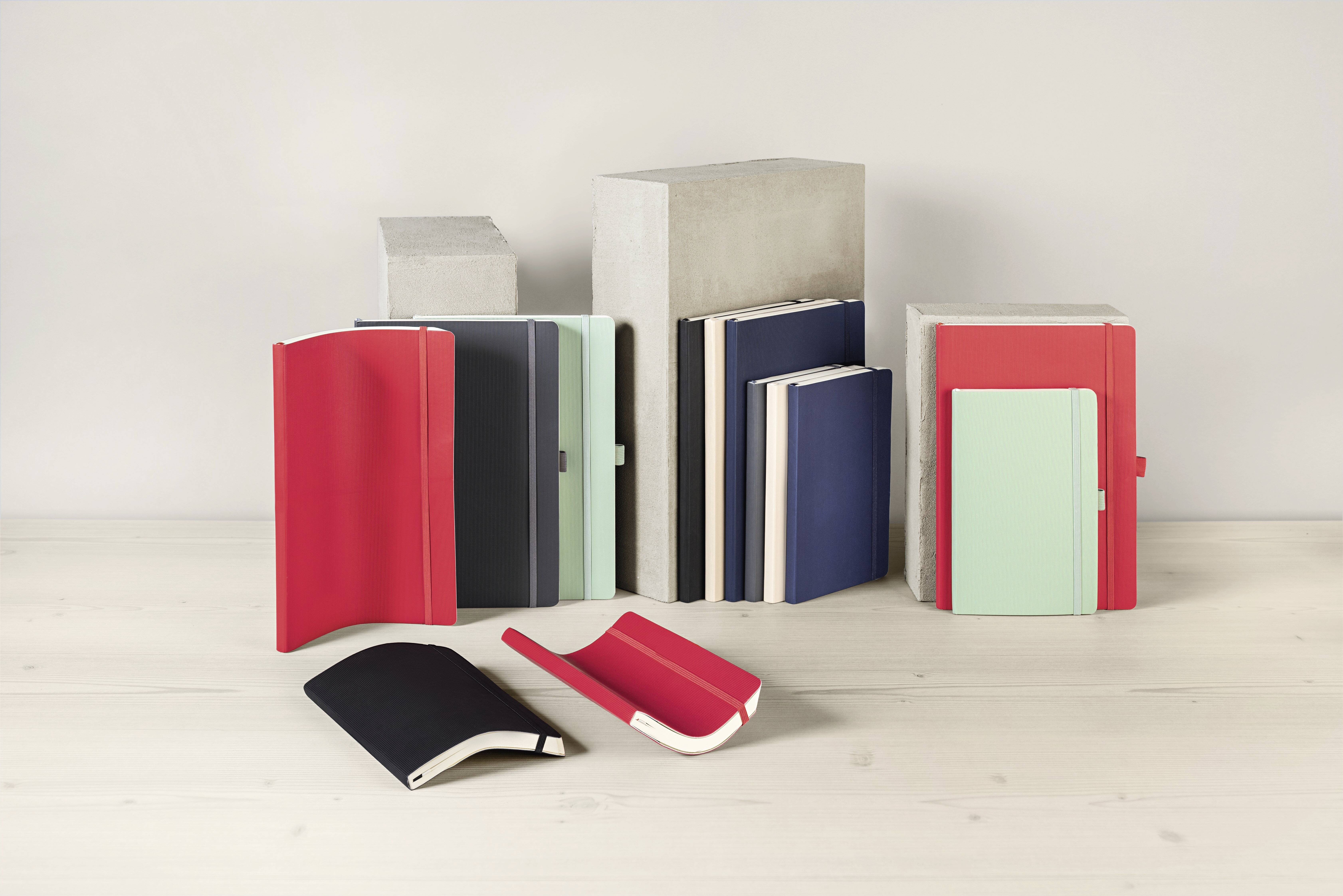 Notizbücher mit roten, schwarzen, blauen und mintfarbenen Einbänden werden stehend und liegend auf einer hellen Holzoberfläche vor grauen Blöcken präsentiert.