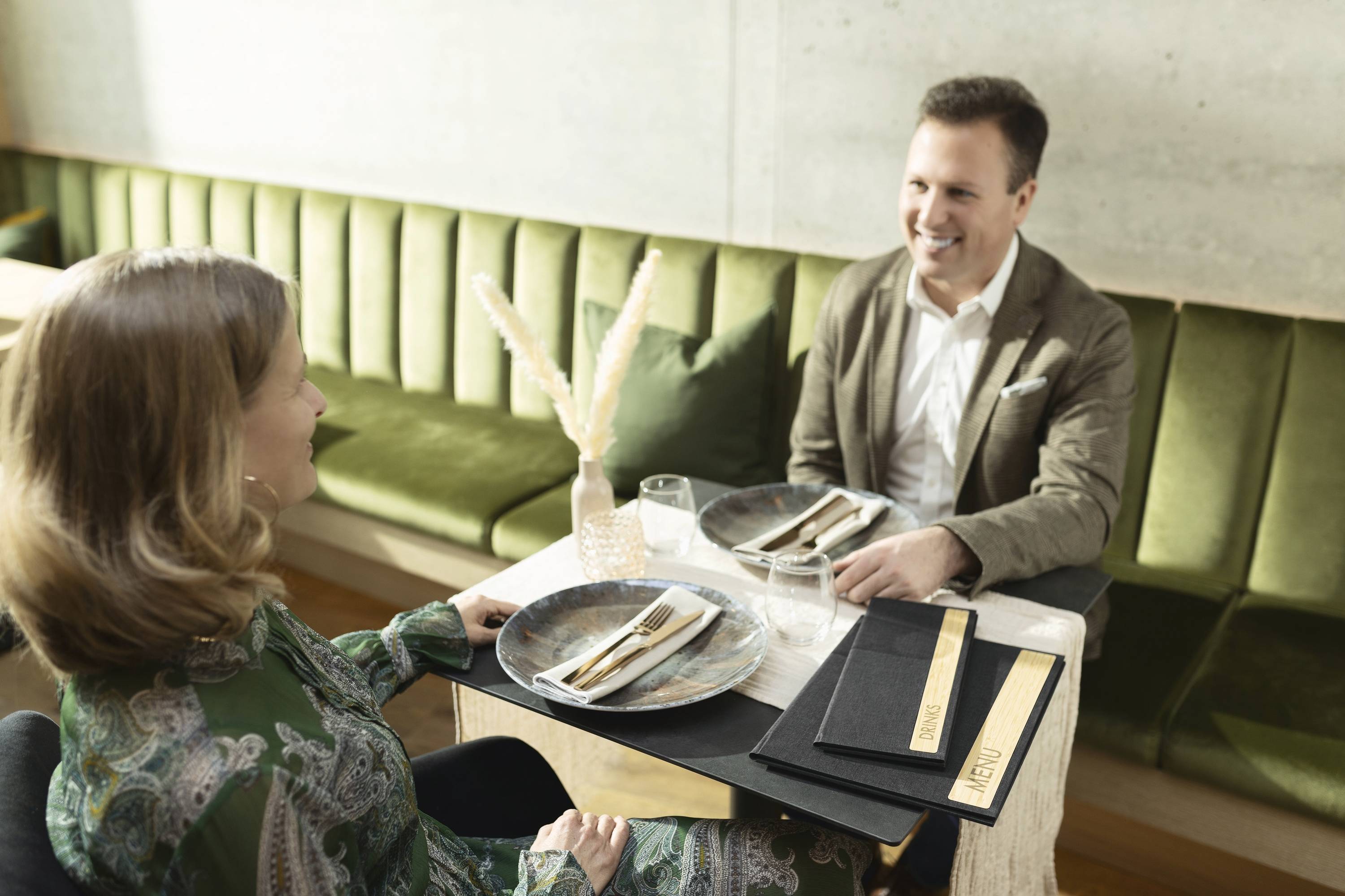 Ein Paar sitzt an einem Tisch in einem Restaurant und lächelt einander an. Der Tisch ist gedeckt mit Tellern, Besteck und Wassergläsern.
