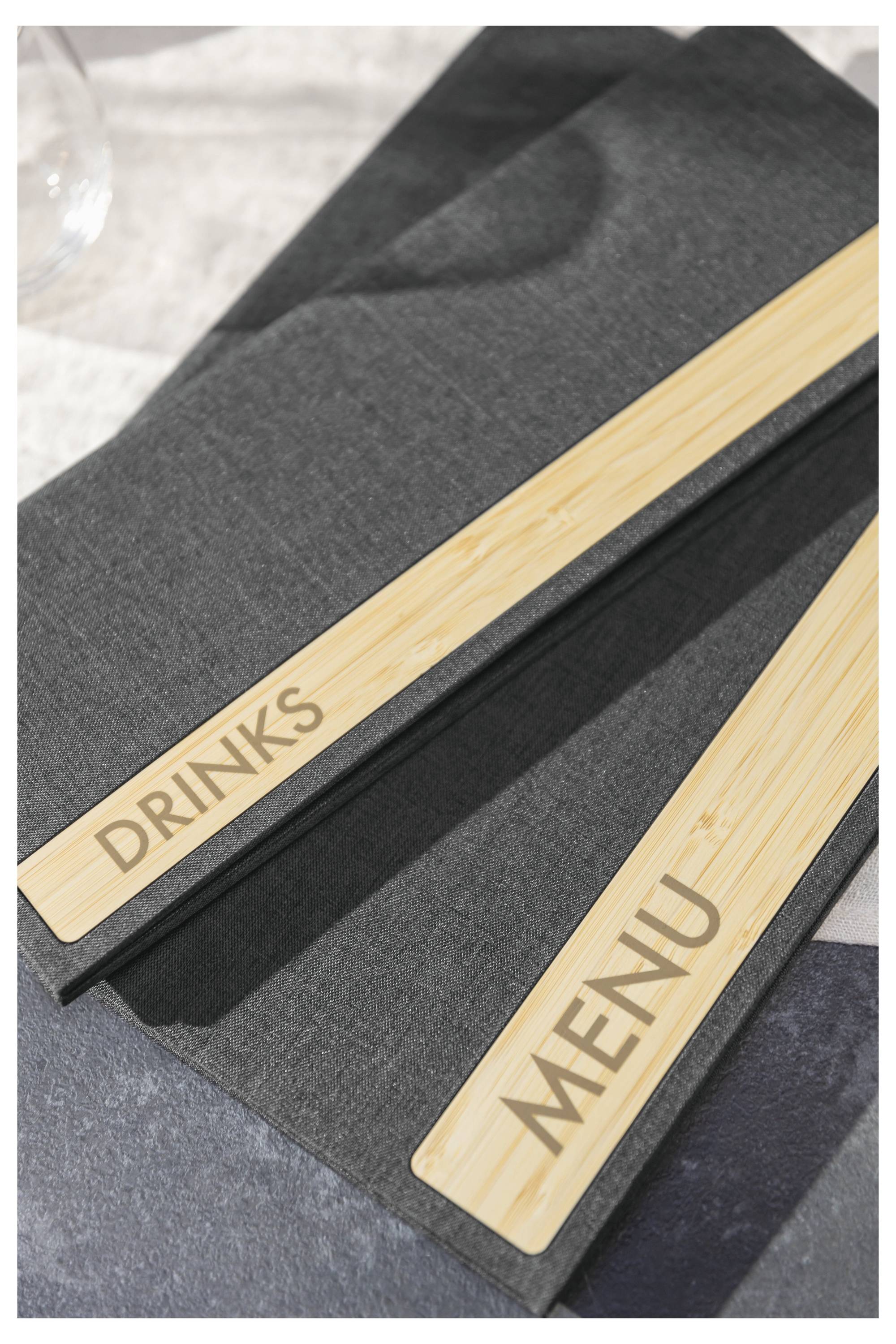 Zwei graue Speisekarten auf einem Tisch, eine mit der Aufschrift ‚GETRÄNKE' und die andere mit ‚SPEISEKARTE', mit Holzleisten auf den Einbänden.