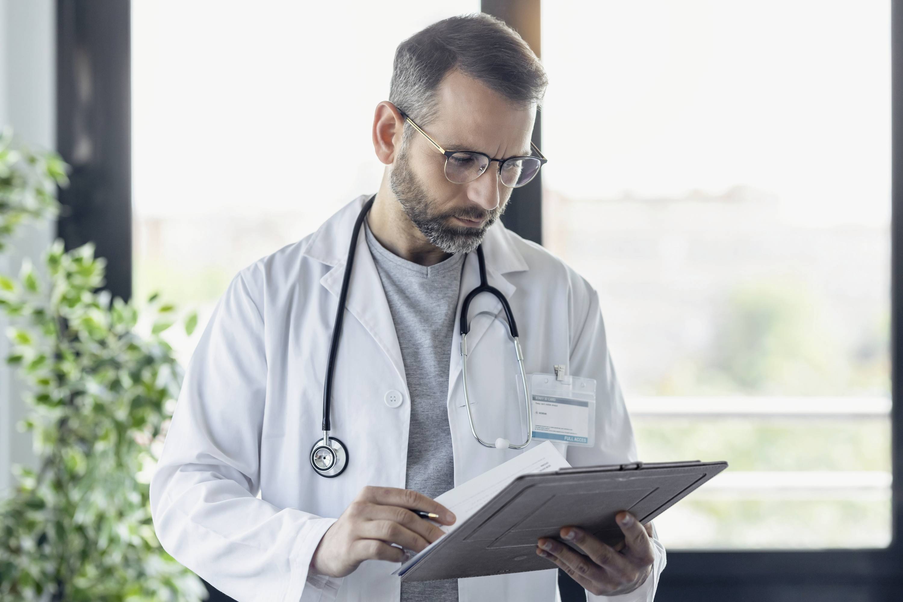 Ein Arzt im Laborkittel, einen Klemmbrett in der Hand, steht neben einem Fenster. Er wirkt konzentriert und scheint möglicherweise medizinische Unterlagen zu prüfen.