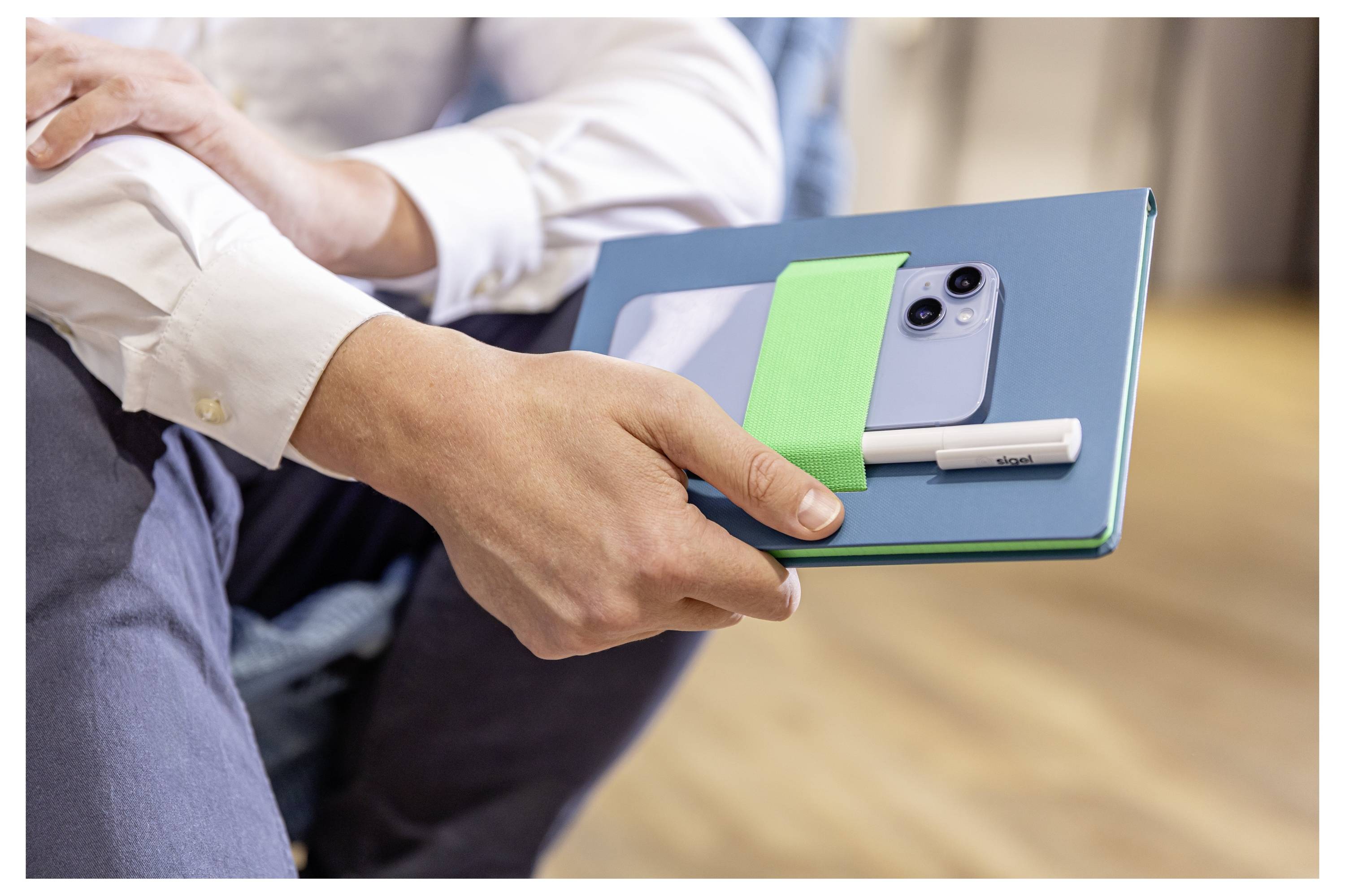 Person, der ein blaues Notizbuch mit einem Smartphone und einem weißen Stift hält, der mit einem grünen Band daran befestigt ist.