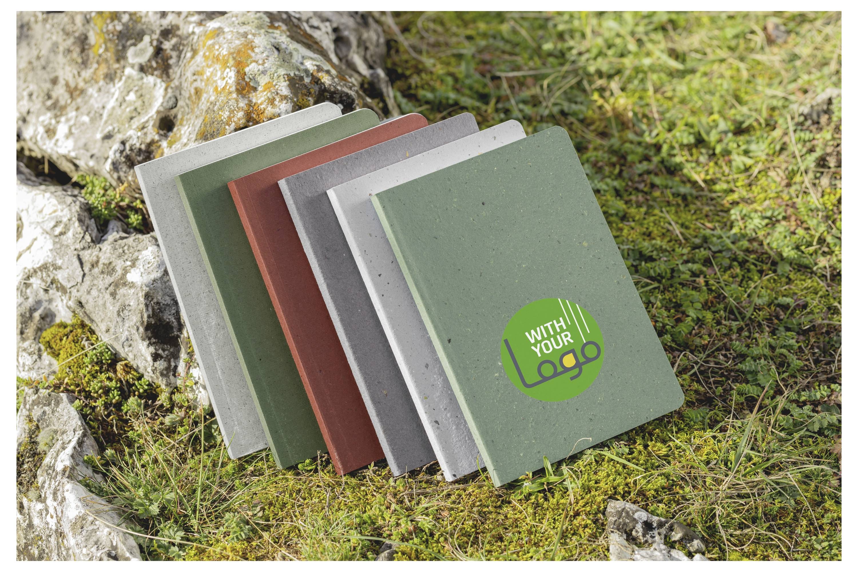 Sieben kleine Notizbücher mit farbigen Umschlägen sind auf Gras neben Steinen fächerartig ausgebreitet, wobei das vordere Notizbuch mit „MIT IHREM LOGO" beschriftet ist.