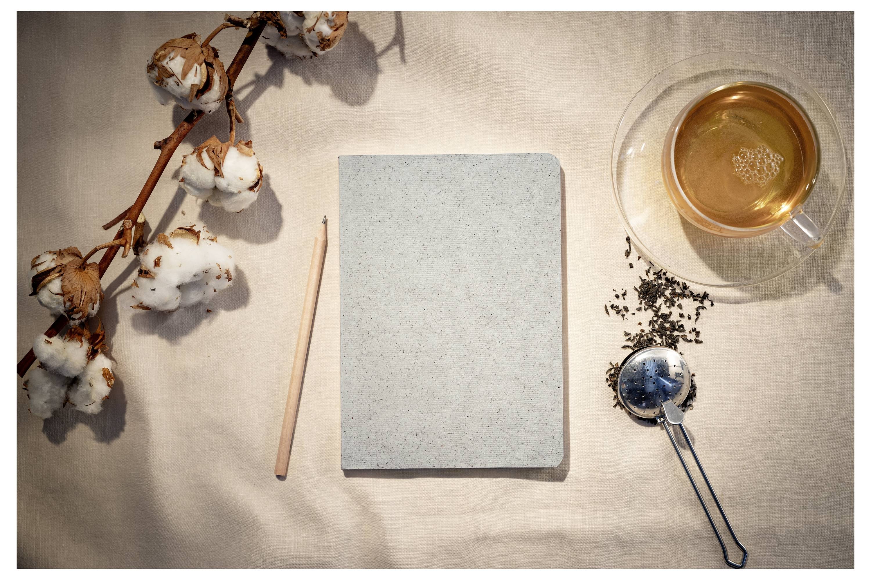Draufsicht eines leeren Notizbuches mit Bleistift, Baumwollzweigen und einer Tasse Tee mit losen Teeblättern und Teefilter auf einem beigefarbenen Tuch.