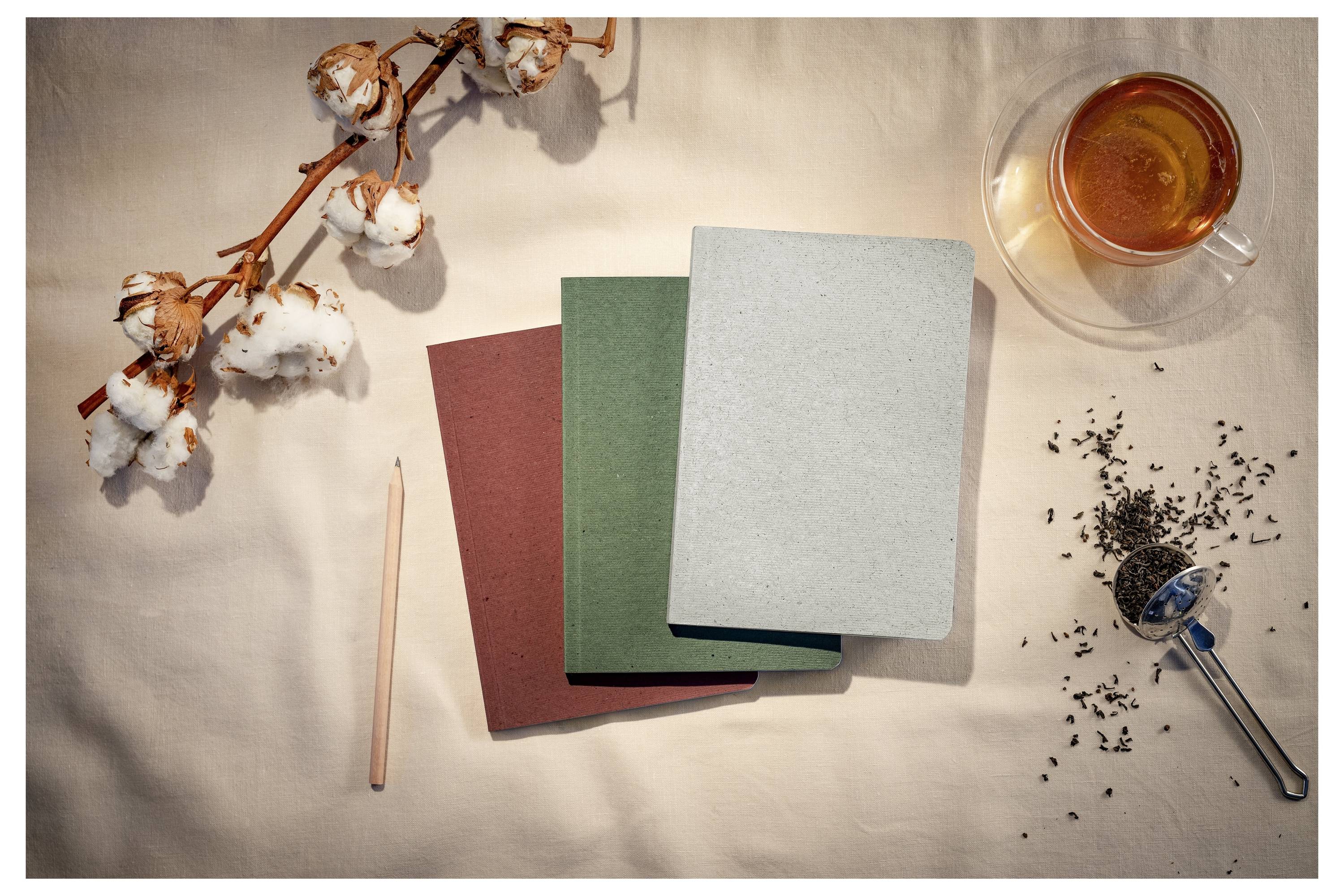 Drei leere Notizbücher, ein Bleistift, Wattestäbchen und eine Tasse Tee auf einer beigen Oberfläche, die ein ruhiges Schreib- oder Tagebuch-Setup suggeriert.