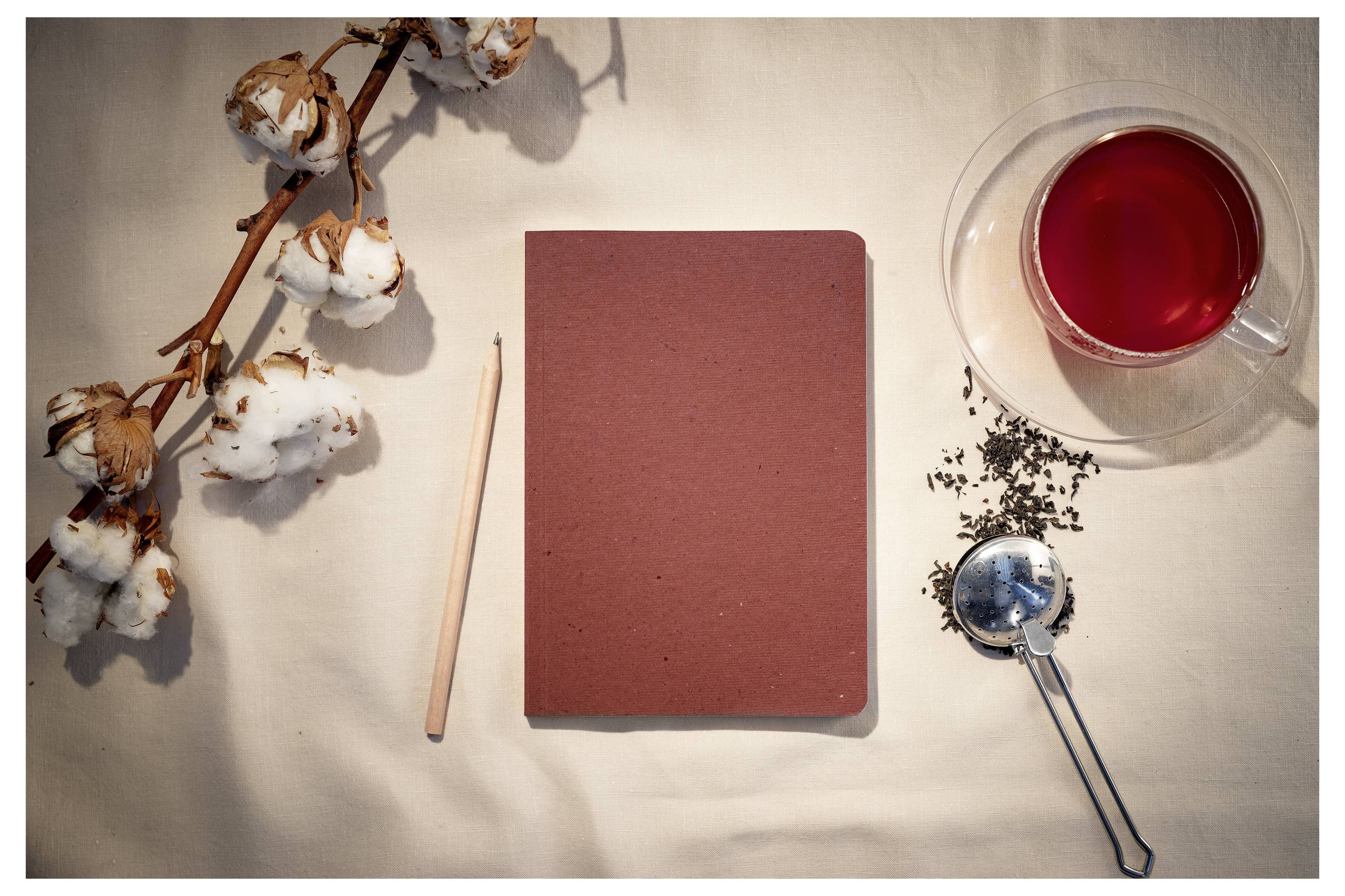 Rotes Notizbuch, zentral neben einem Bleistift, Wattestäbchen, losen Teeblättern und einer Tasse rotem Tee auf beigefarbenem Stoffhintergrund angeordnet.