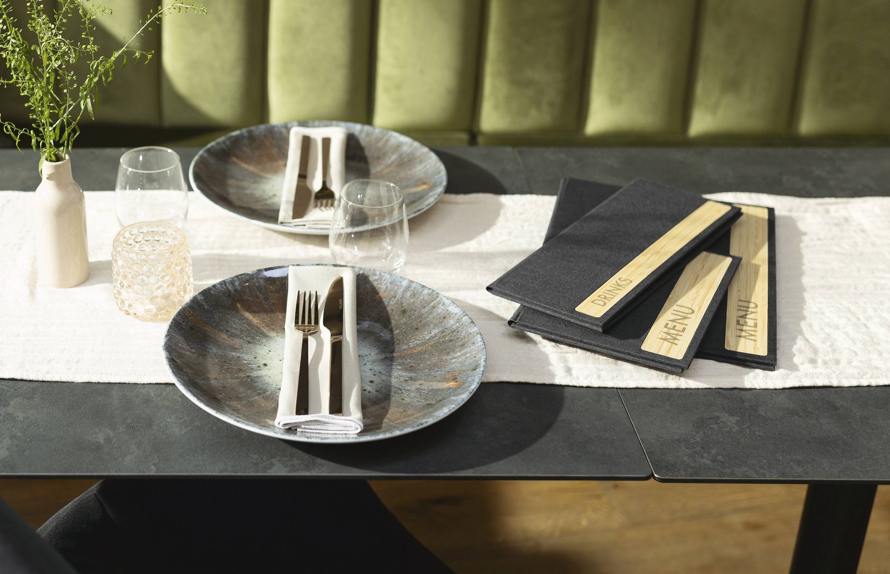 Ein Restauranttisch für zwei Personen, gedeckt mit Keramiktellern, Besteck in Servietten gewickelt und Speisekarten rechts daneben. Eine kleine Vase enthält grüne Zweige.