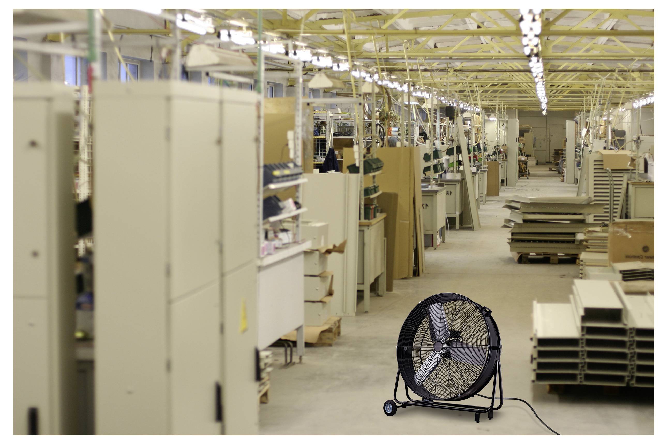 Ein großer Industrieventilator steht auf dem Boden einer geräumigen Fabrikhalle, umgeben von hohen Maschinen und Regalen, die mit Materialien gefüllt sind.