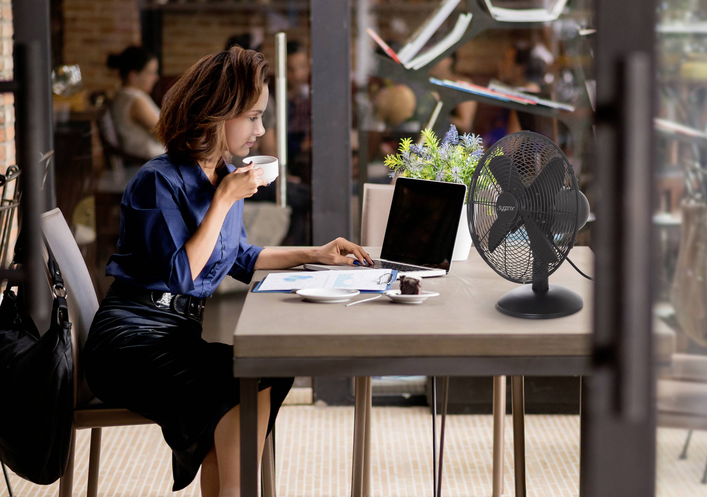 Eine Frau sitzt an einem Cafétisch, trinkt Kaffee und arbeitet an einem Laptop. Ein Ventilator und eine kleine Pflanze stehen auf dem Tisch. Drinnen, entspannte Atmosphäre.