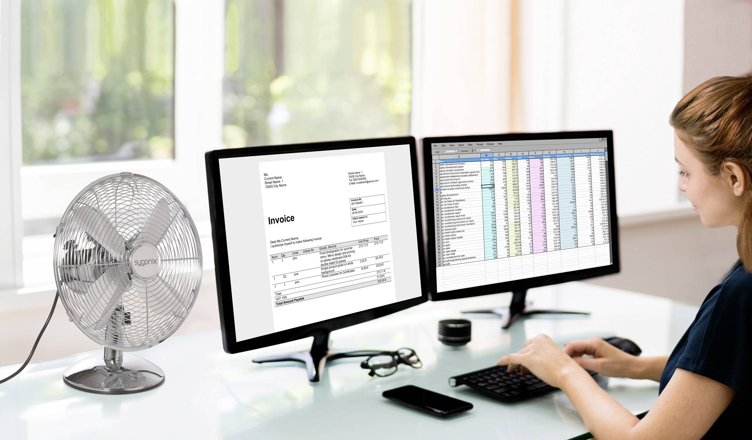 Eine Person arbeitet an einem Schreibtisch mit zwei Computermonitoren, auf denen eine Rechnung und eine Tabellenkalkulation zu sehen sind, neben einem silbernen Schreibtischventilator.
