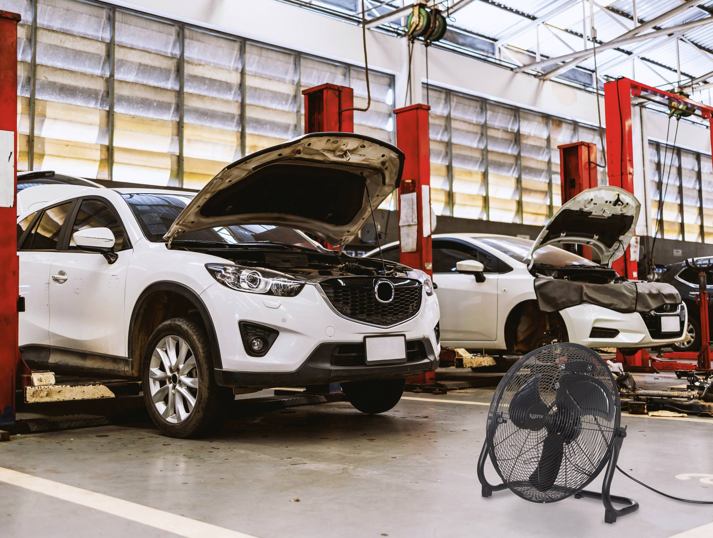 Zwei Autos mit offenen Motorhauben auf hydraulischen Hebebühnen in einer Autowerkstatt; ein großer Ventilator befindet sich im Vordergrund.