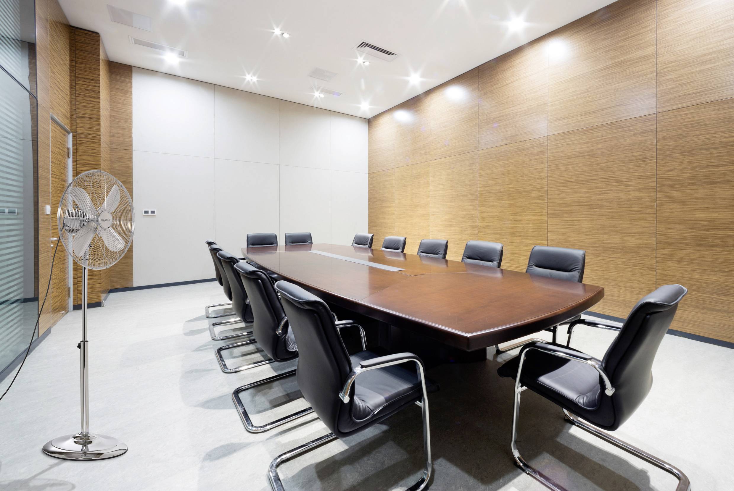 Ein großer Konferenzraum mit einem langen Holztisch, der von zehn schwarzen Stühlen umgeben ist. Der Raum verfügt über einen Ventilator, Holzvertäfelungswände und helle Beleuchtung.
