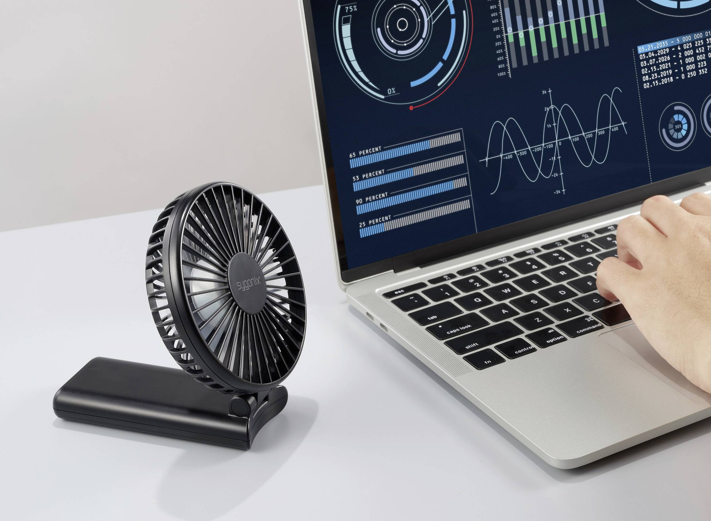 Ein tragbarer Ventilator, der neben einem Laptop mit analytischen Grafiken auf dem Bildschirm platziert ist; eine Hand tippt auf der Tastatur des Laptops und deutet auf eine Arbeitsumgebung hin.