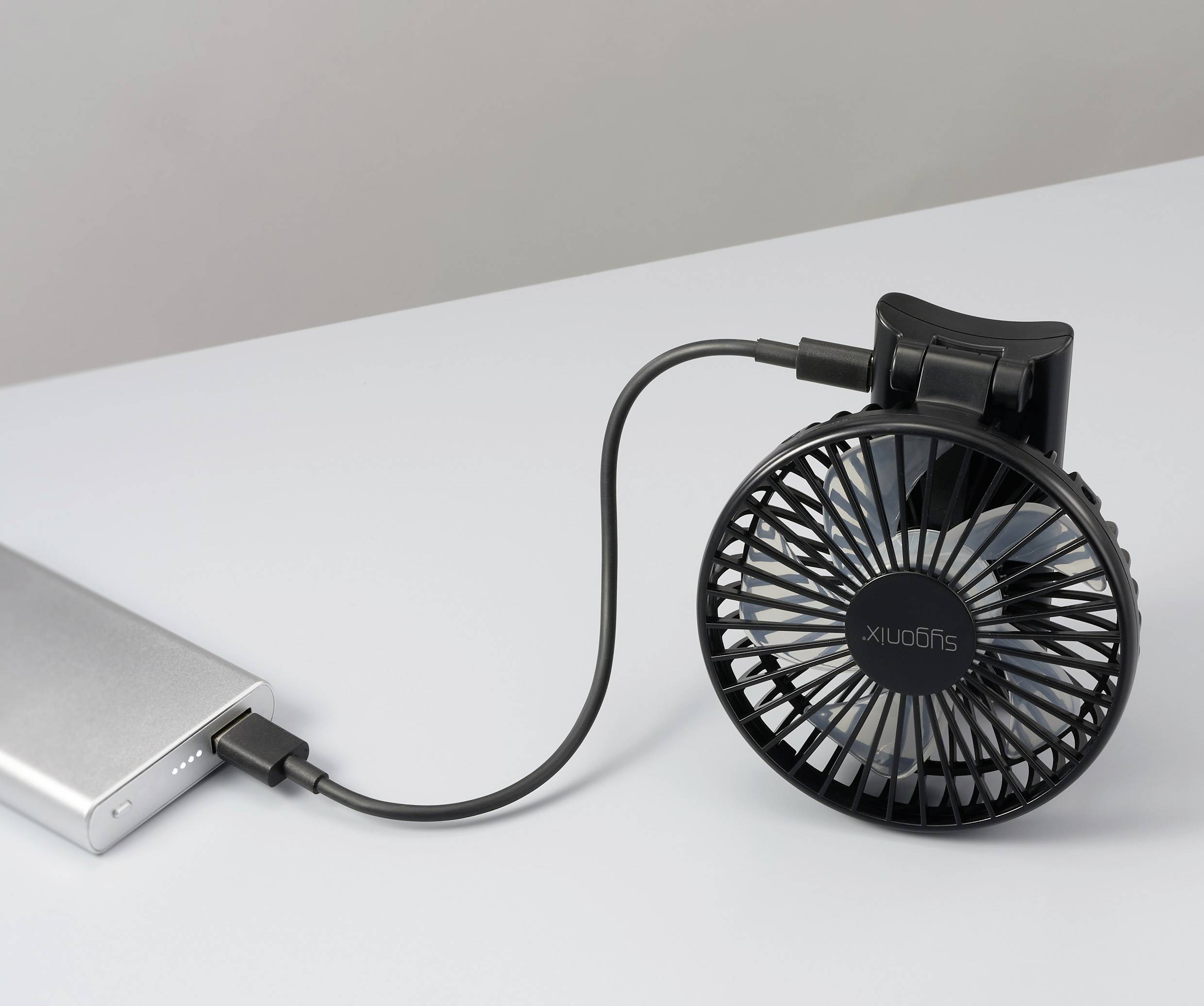 Ein kleiner schwarzer Ventilator, der über ein USB-Kabel mit einer tragbaren Powerbank auf einer weißen Oberfläche verbunden ist und seine Mobilität und Benutzerfreundlichkeit demonstriert.