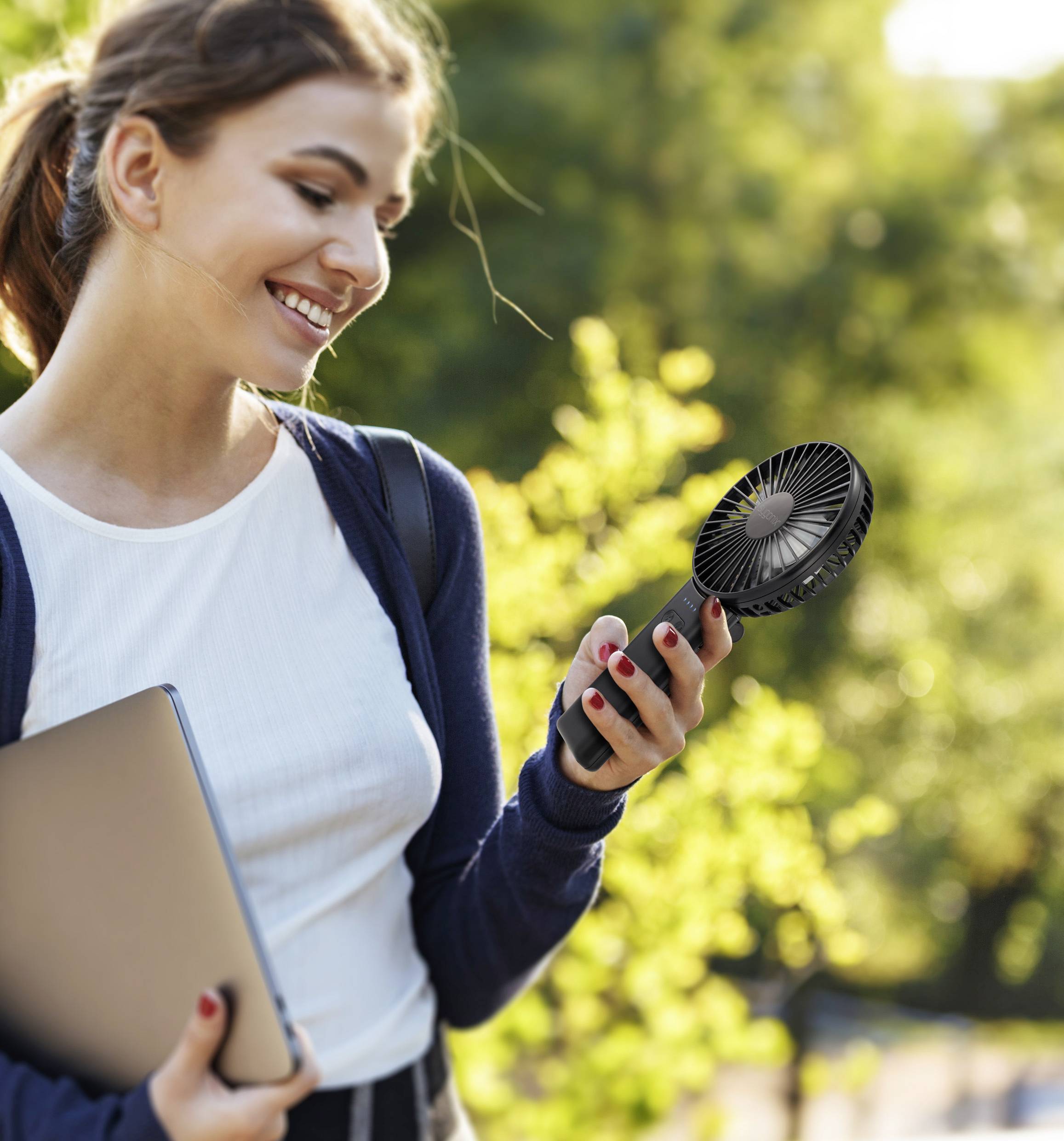 Eine Person lächelt, während sie einen tragbaren Ventilator hält und einen Laptop in einem sonnigen, grünen Parkgelände trägt.