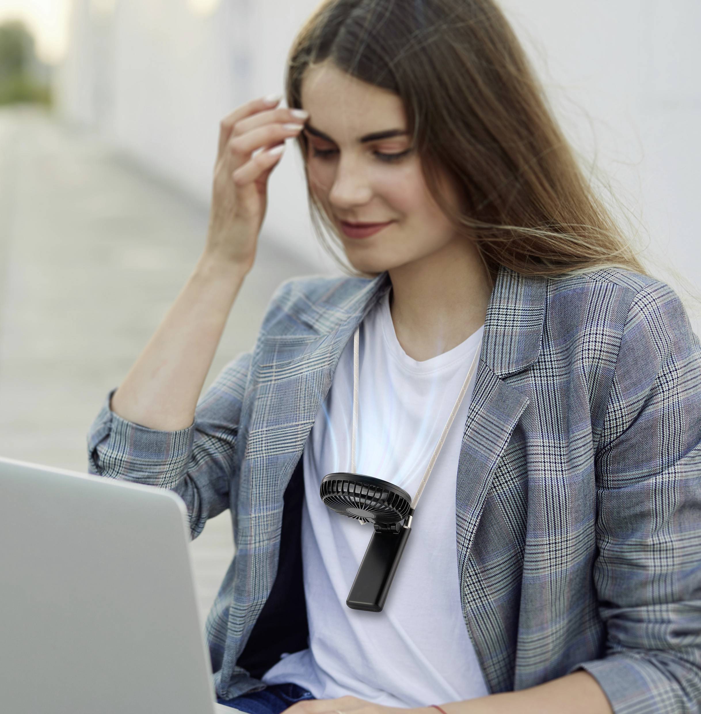Eine Frau sitzt im Freien und arbeitet an einem Laptop, trägt einen tragbaren Nackenlüfter und einen Karosakko, was auf eine lockere Arbeitsumgebung an einem warmen Tag hindeutet.