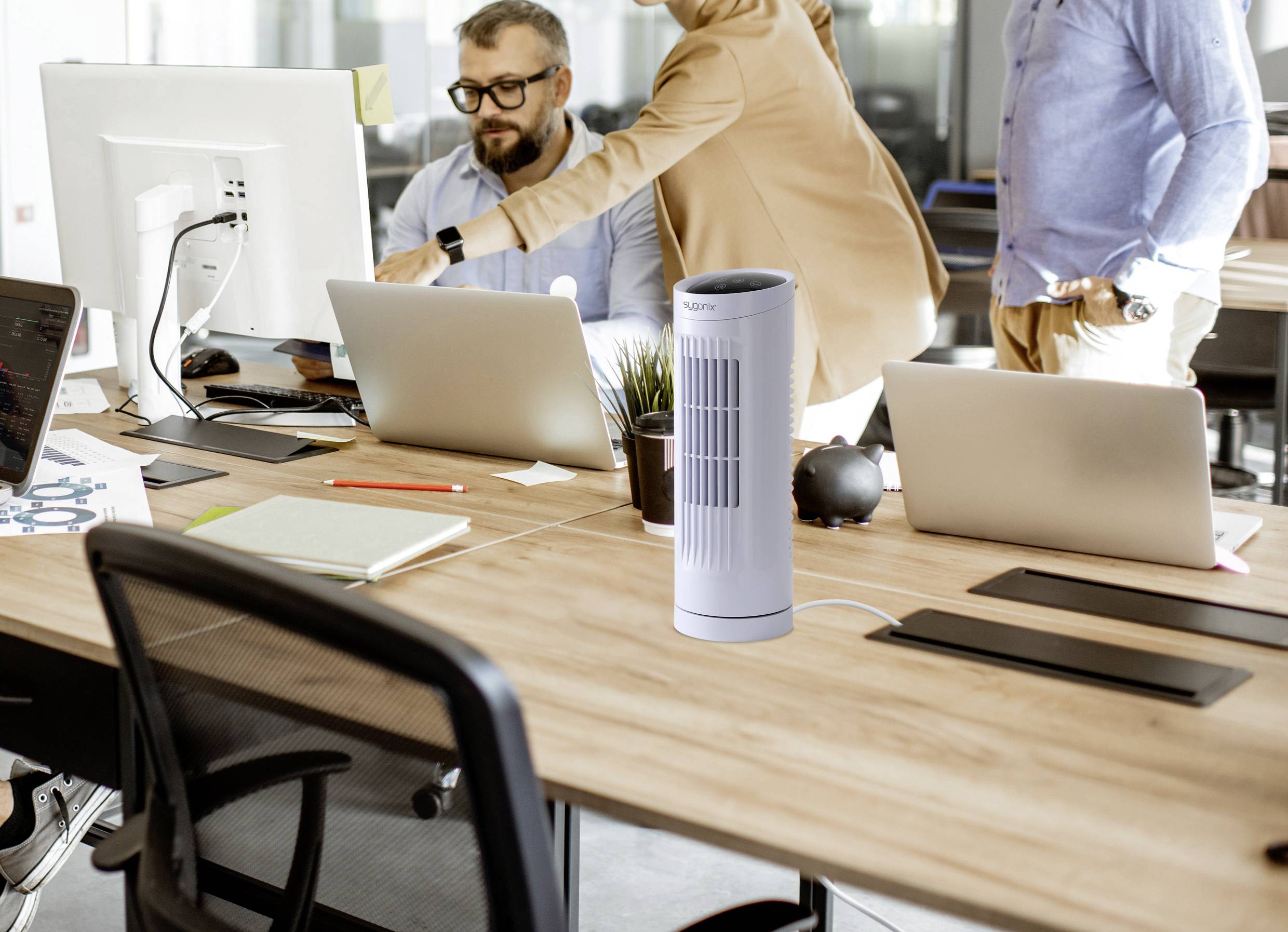 Eine Büroumgebung mit drei Personen, die an Schreibtischen mit Computern arbeiten. Ein Turmventilator steht im Vordergrund auf einem Schreibtisch.