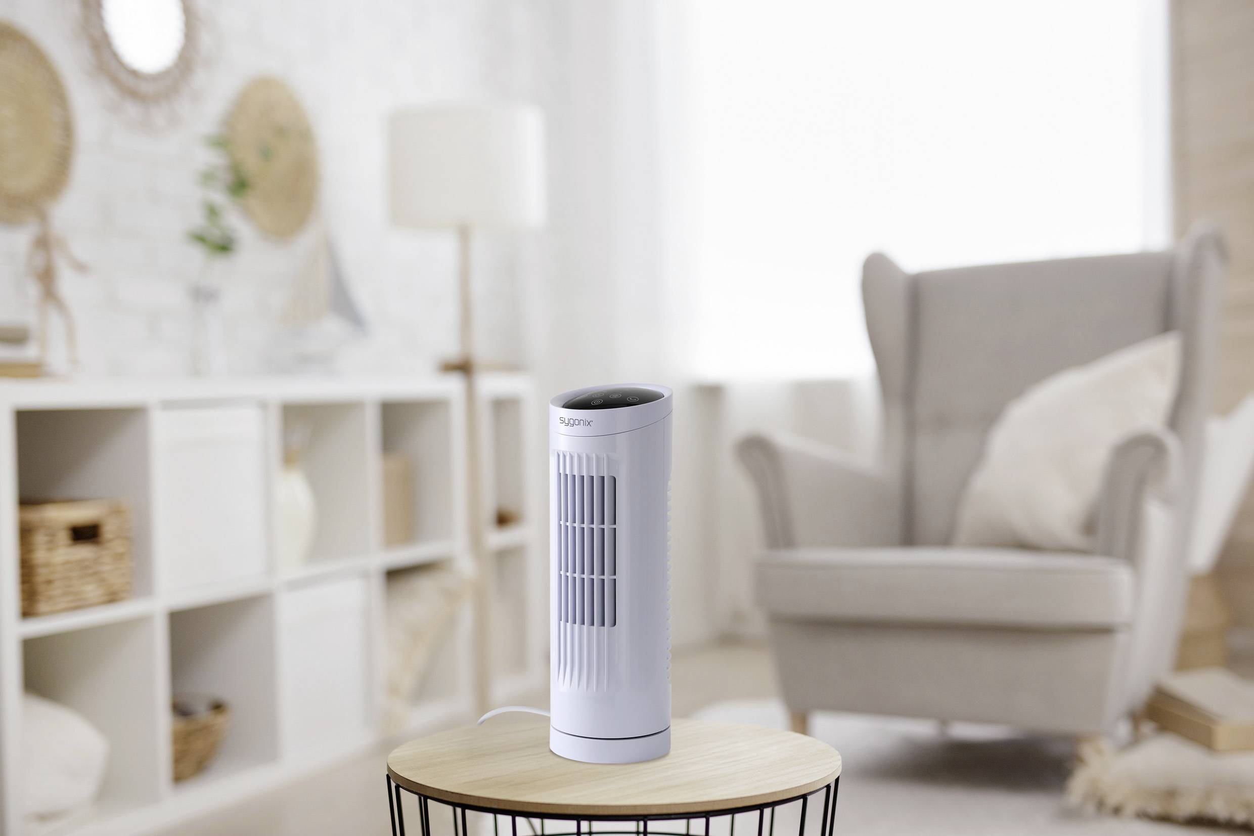 Ein kleiner, weißer Turmventilator auf einem runden Tisch in einem hellen, gemütlichen Wohnzimmer mit einem grauen Sessel und dekorativen Regalen im Hintergrund.